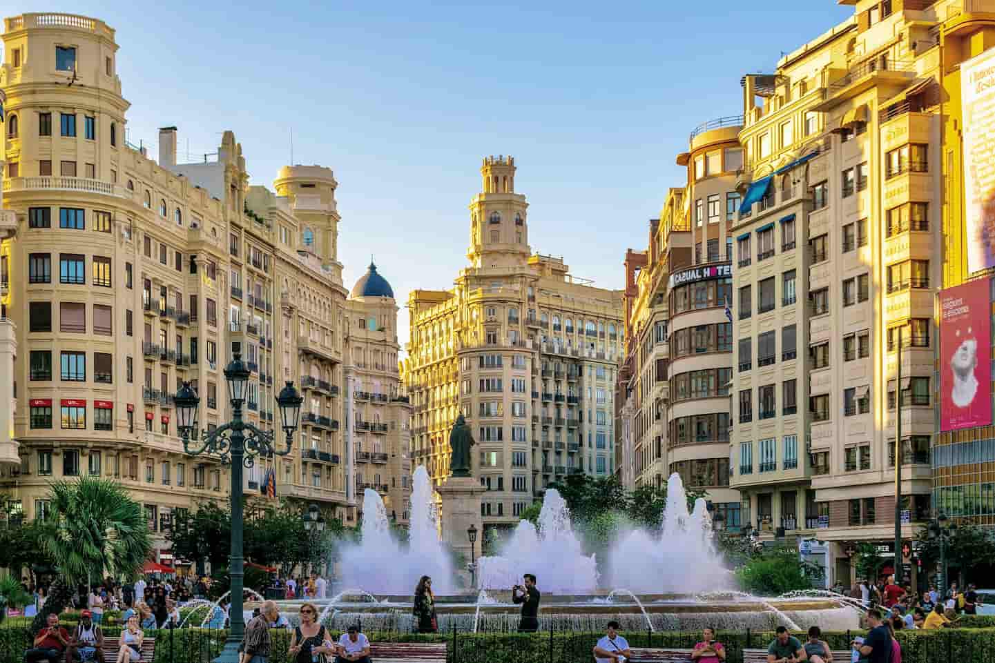 An urban plaza with people strolling, ornate buildings under a clear sky, and a large fountain with multiple jets in the center.