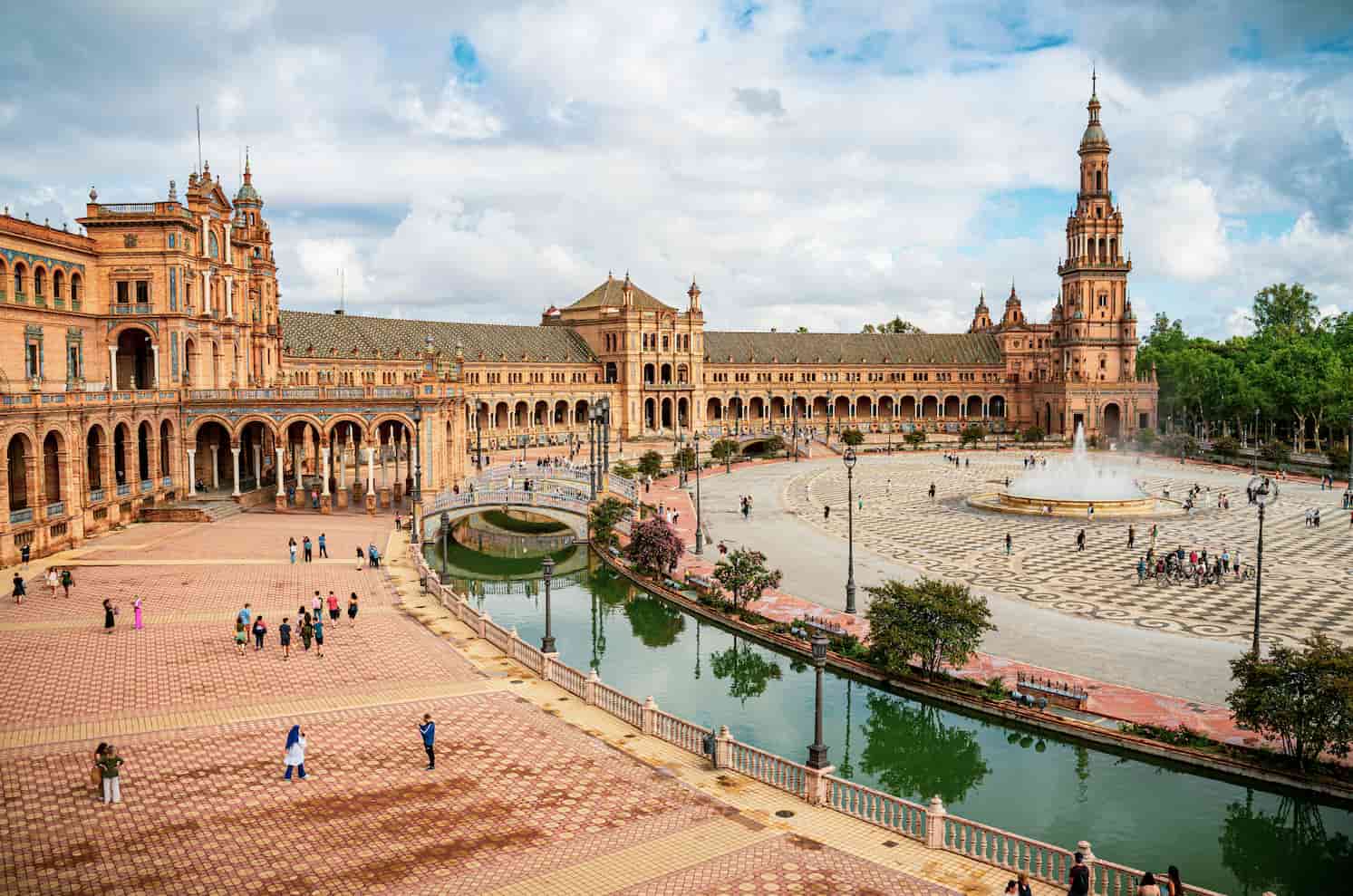 A wide plaza with a semi-circular building, arched colonnade, tower, canal, and fountain, surrounded by people under a partly cloudy sky.