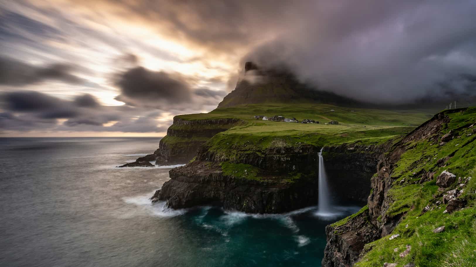 Dramatic coastline with a waterfall cascading over green cliffs into the ocean, under a cloudy sky with a mountain in the background.