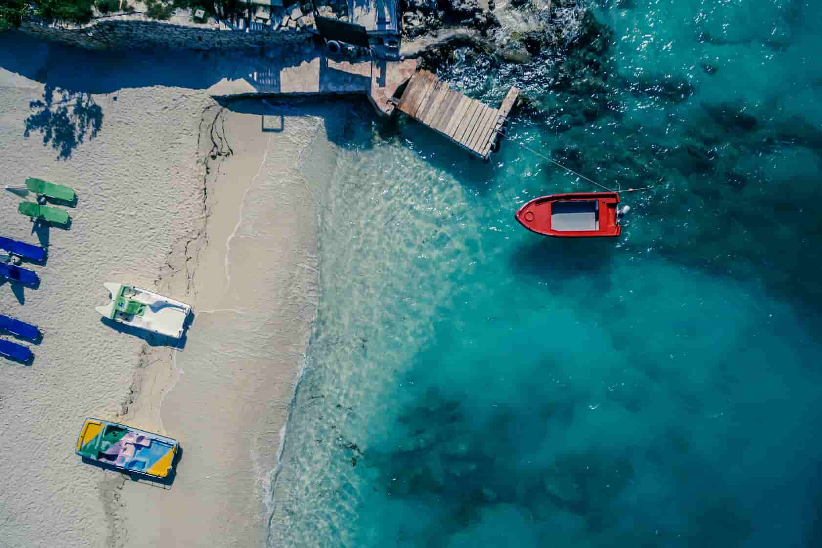 Aerial view of a sandy beach with sun loungers and boats in clear blue water. A red boat is near a wooden dock by the shoreline.