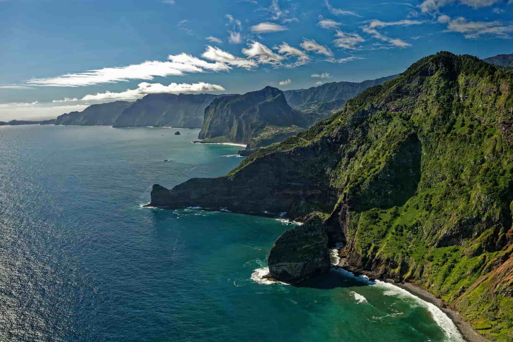 Aerial view of a rugged coastline with steep green cliffs, clear blue ocean, and scattered white clouds in a bright blue sky.