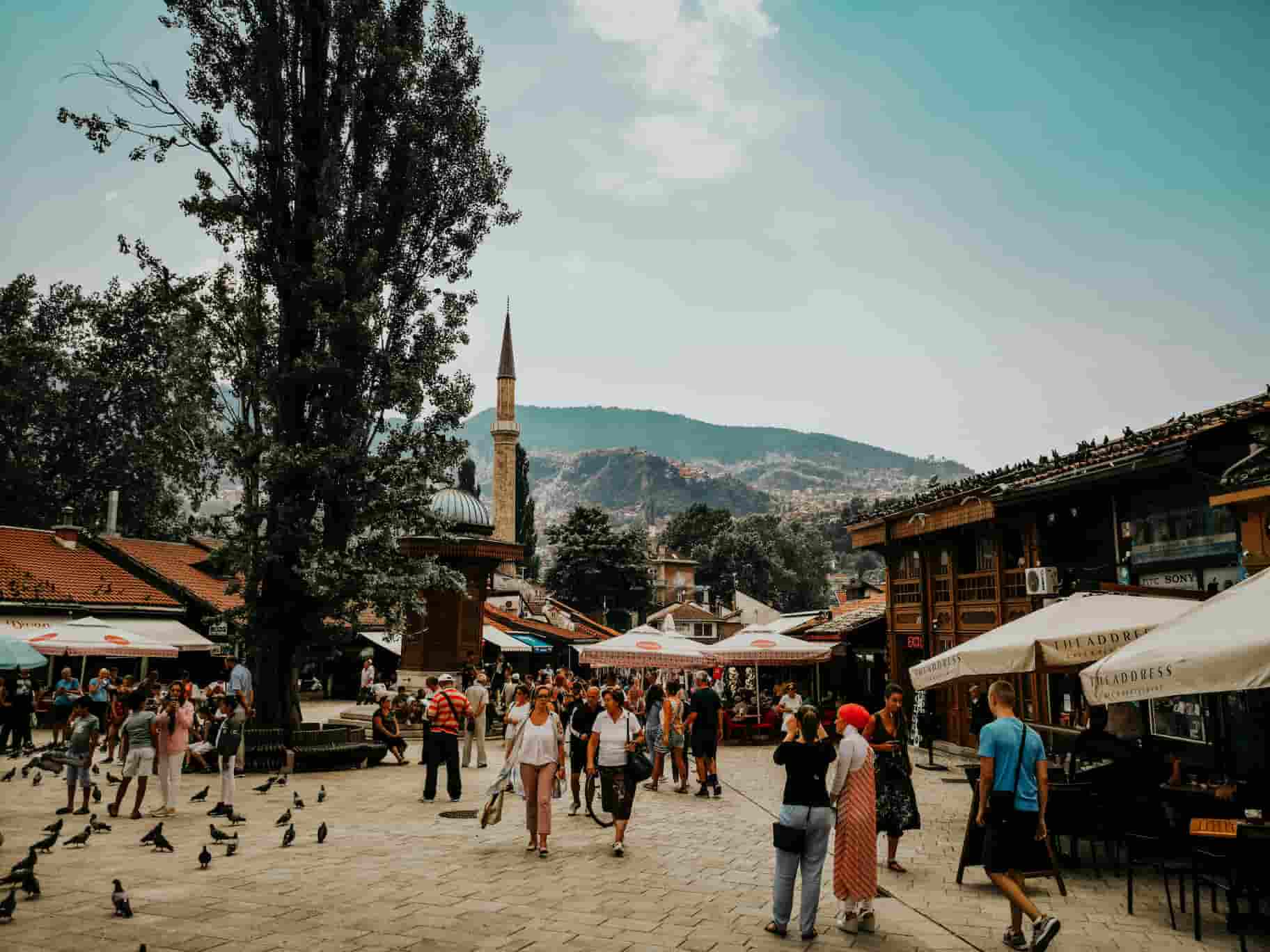 Bustling square with people, pigeons, outdoor cafes, and historic buildings. A tall minaret is visible against a backdrop of hills and a clear sky.