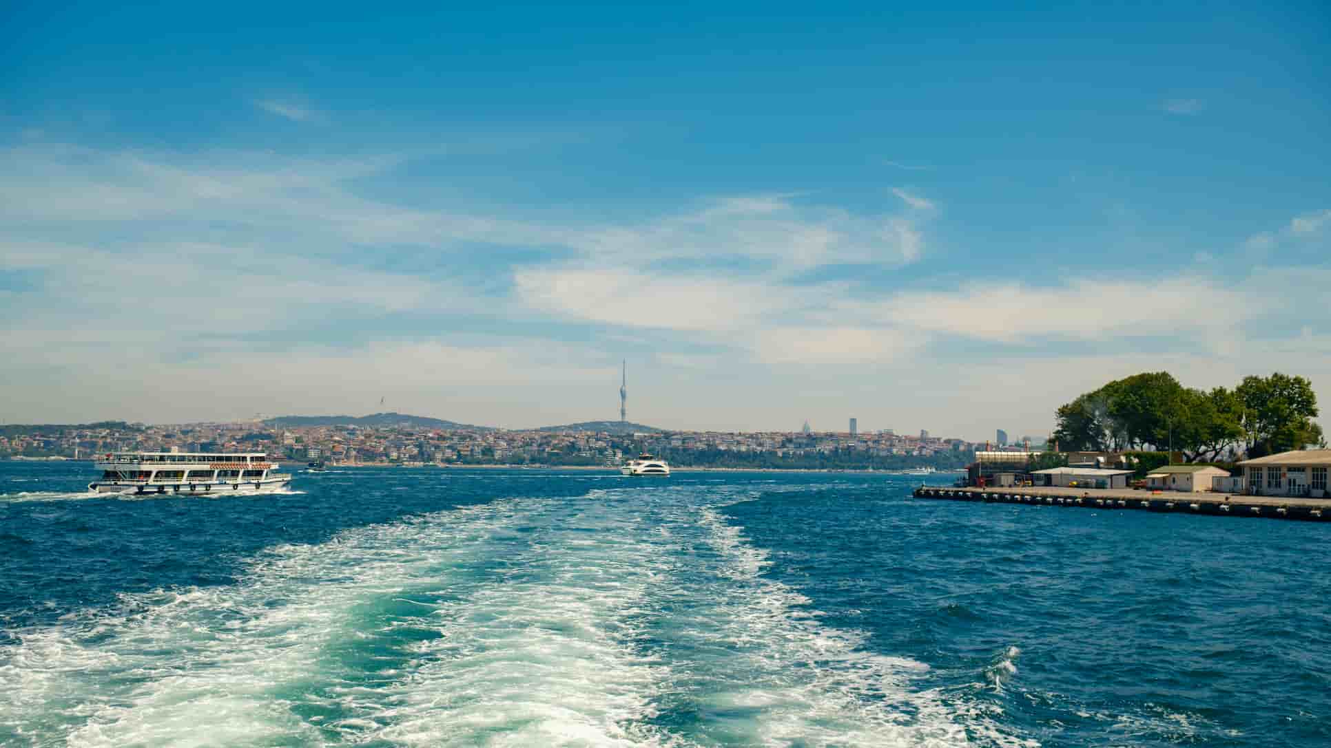 View of a city skyline and tower in the distance, with two boats on a blue sea and a pier with trees to the right, under a clear blue sky.