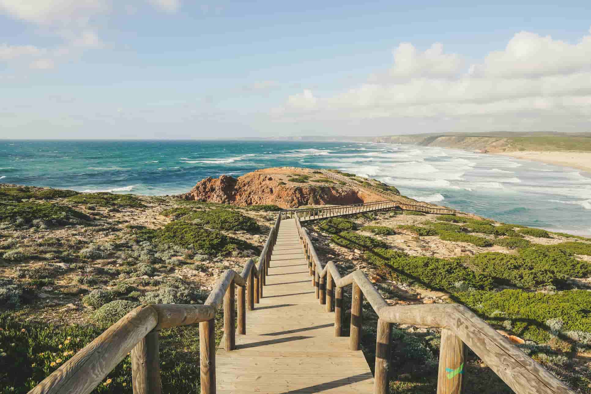 Wooden walkway leading to a scenic coastal view with rocky cliffs, lush greenery, and waves crashing on a sandy beach under a partly cloudy sky.