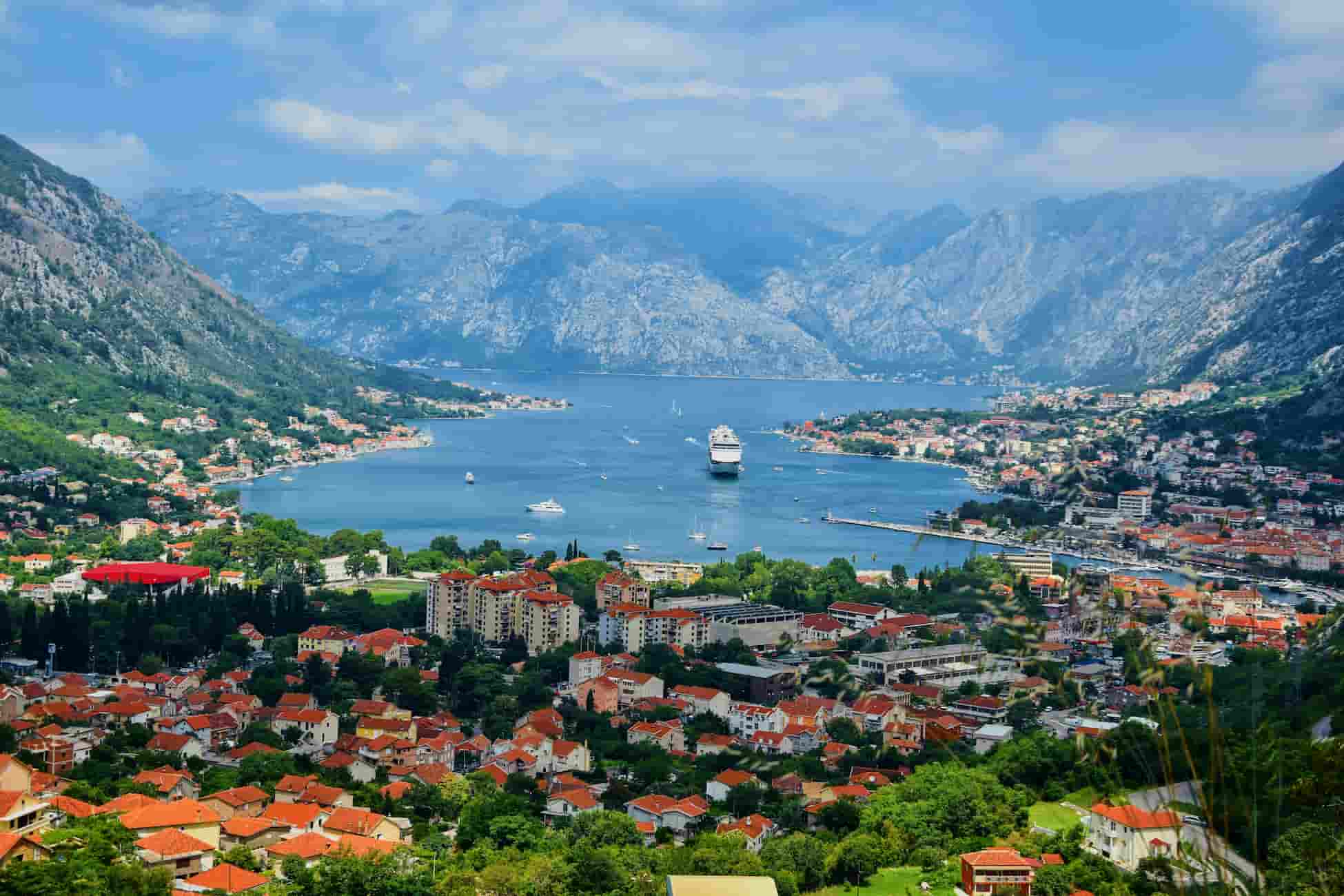 Scenic view of Kotor Bay in Montenegro, featuring a cruise ship on the water, surrounded by mountainous landscape and red-roofed buildings.