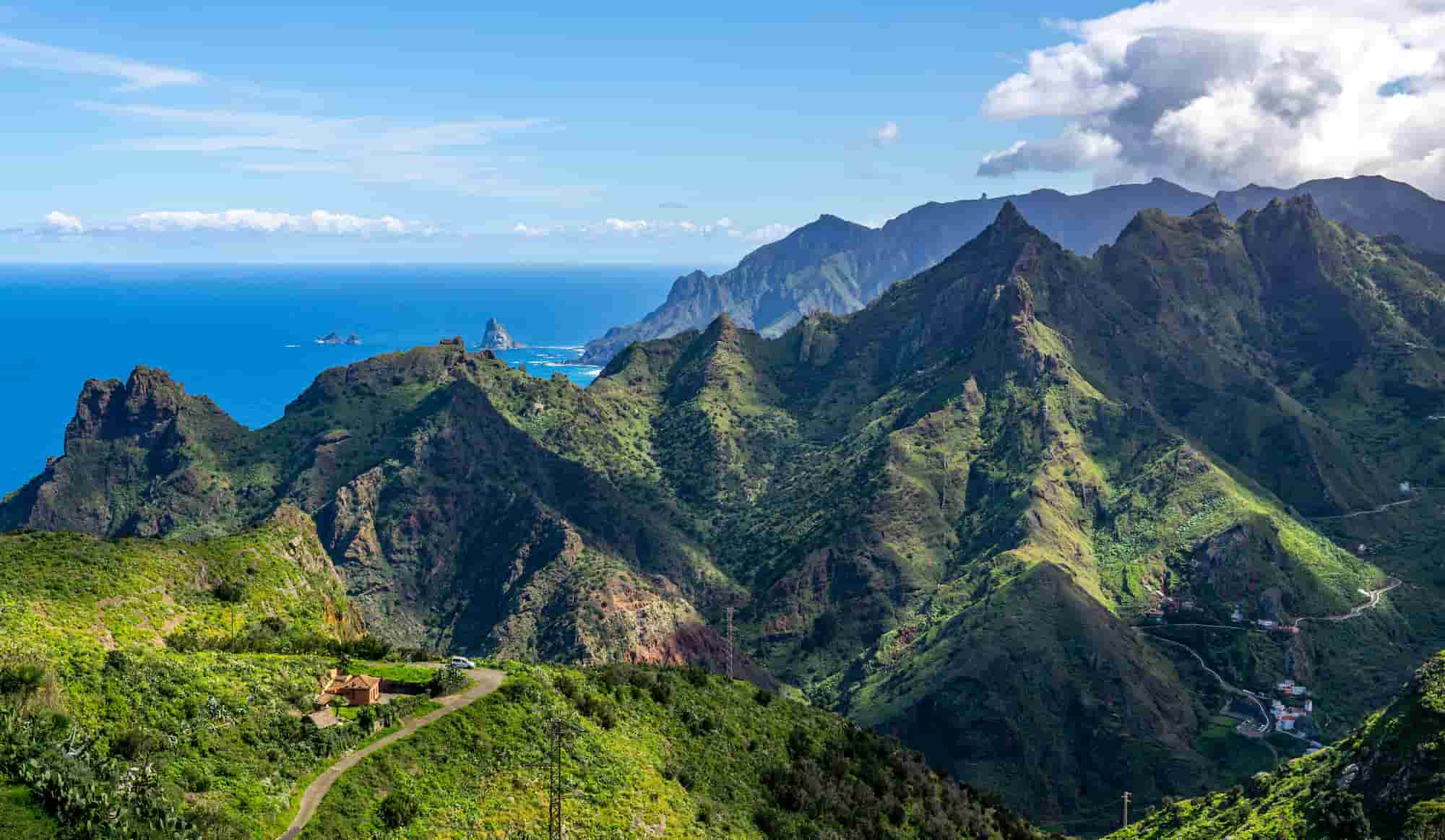 Lush green mountains under a bright blue sky with distant ocean views, dotted with small houses and winding roads.
