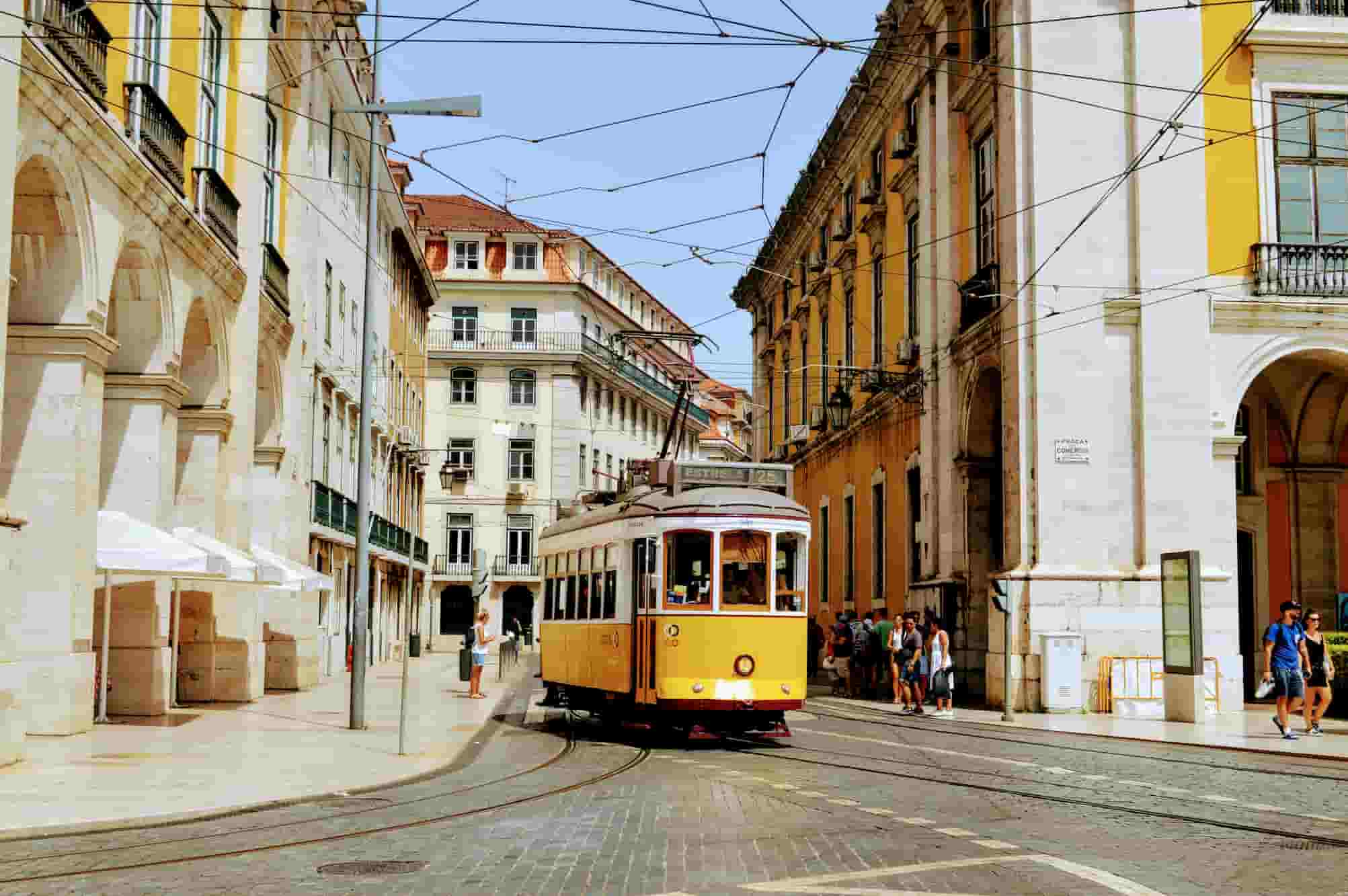 Vintage yellow tram on a street in Lisbon, surrounded by historic buildings, with overhead wires and people walking nearby.