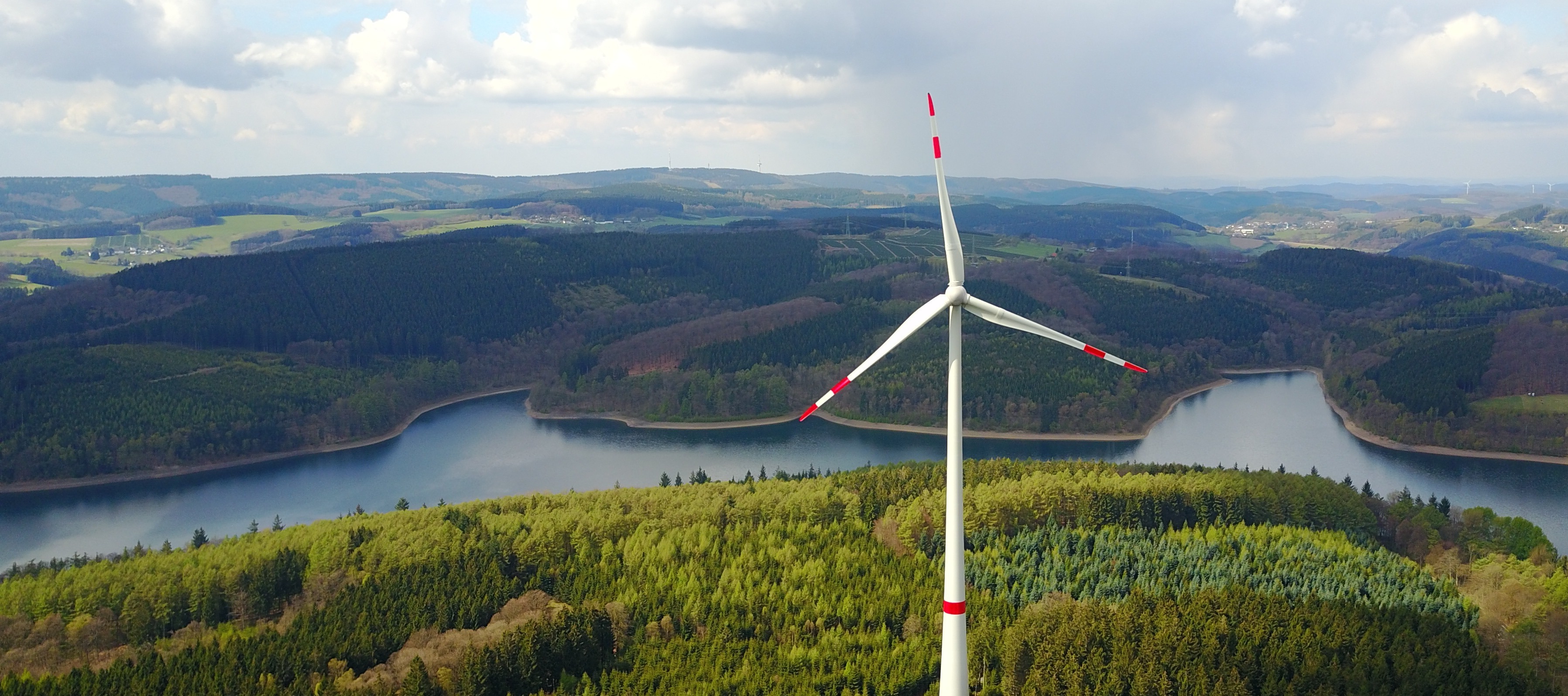 Windkraftwerk im Wald vor einem Fluss mit Bergen im Hintergrund