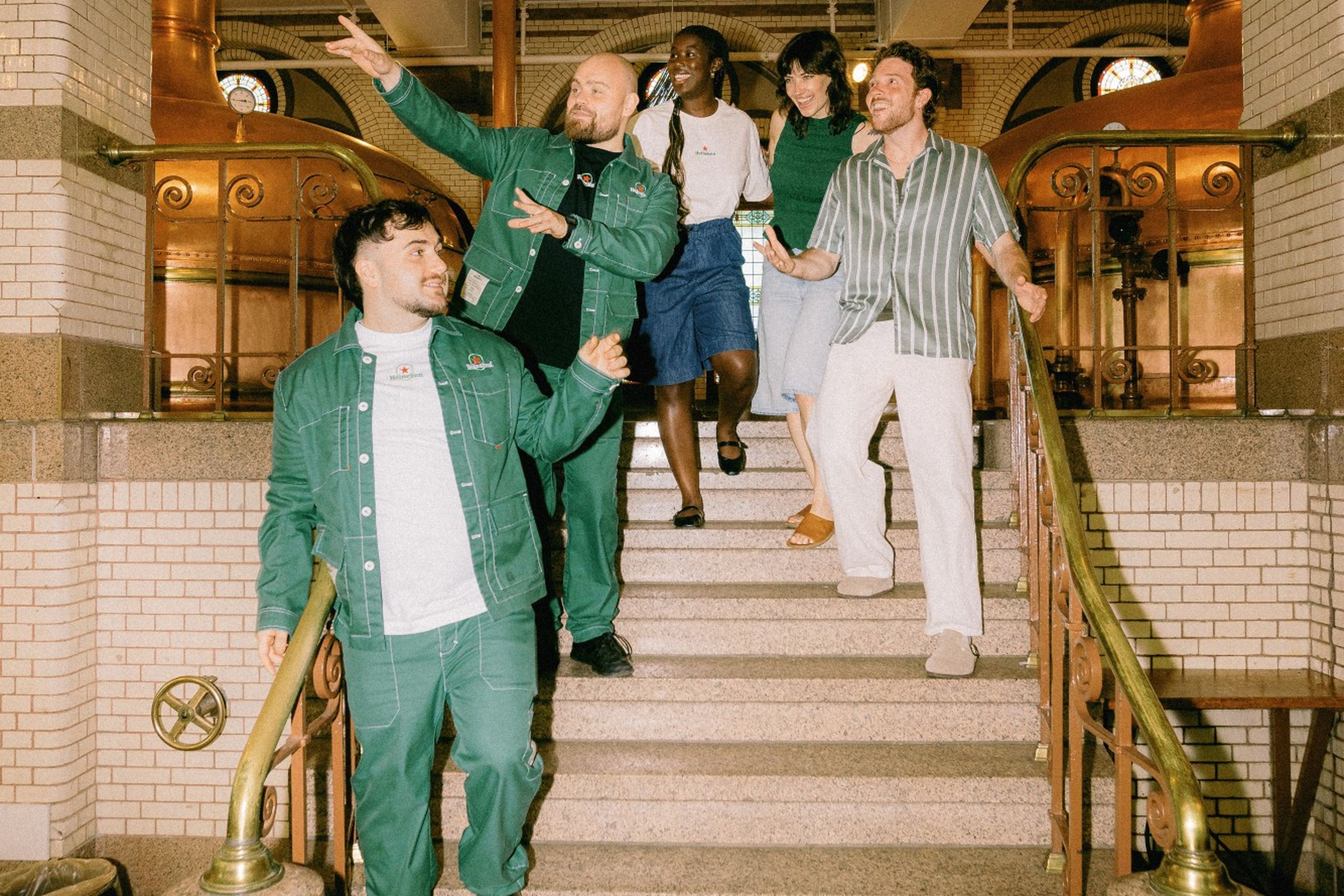 Five cheerful friends walk down grand stairs in an ornate building with copper fixtures, white tiles, and brass railings.