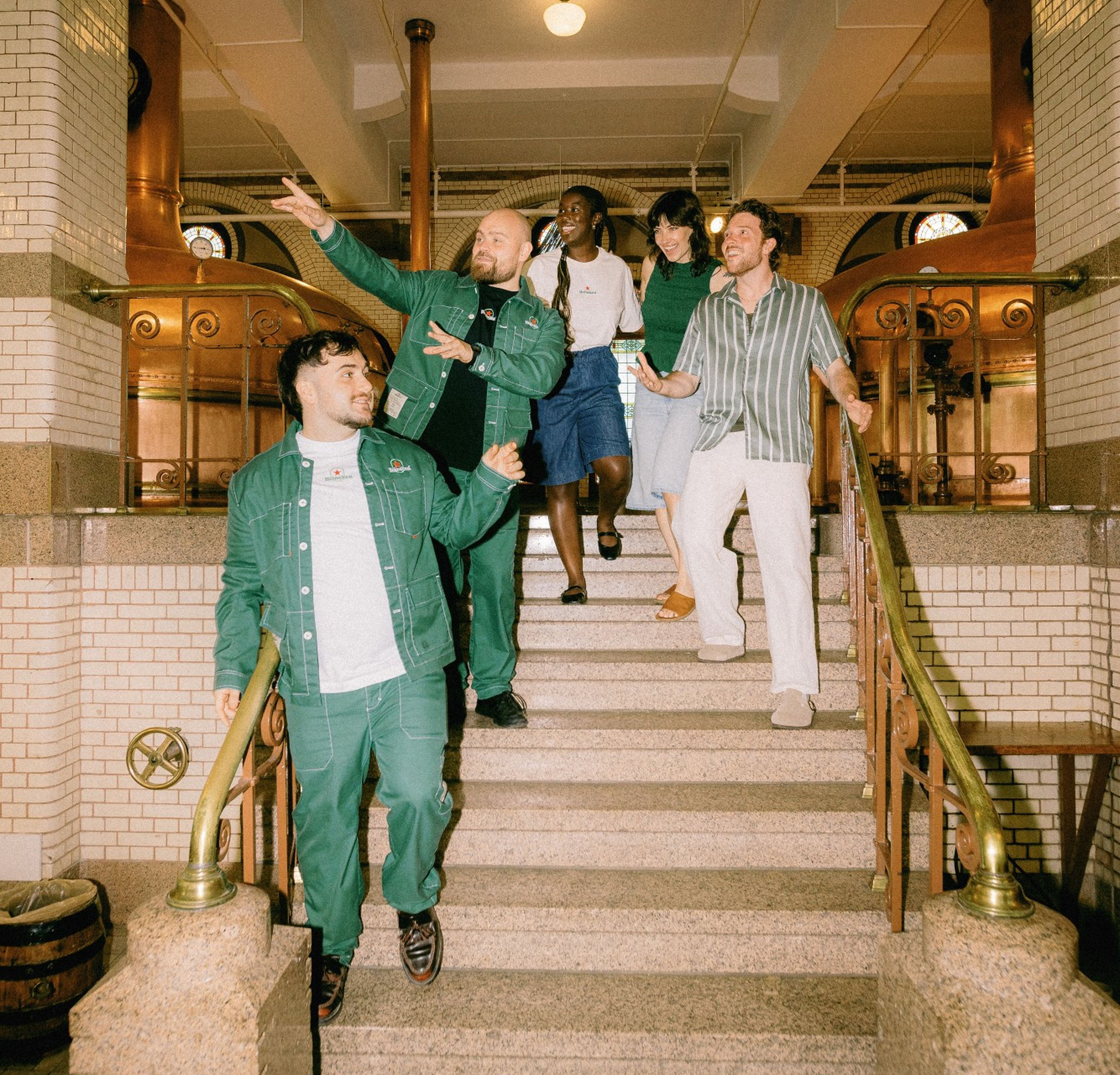Five cheerful friends walk down grand stairs in an ornate building with copper fixtures, white tiles, and brass railings.