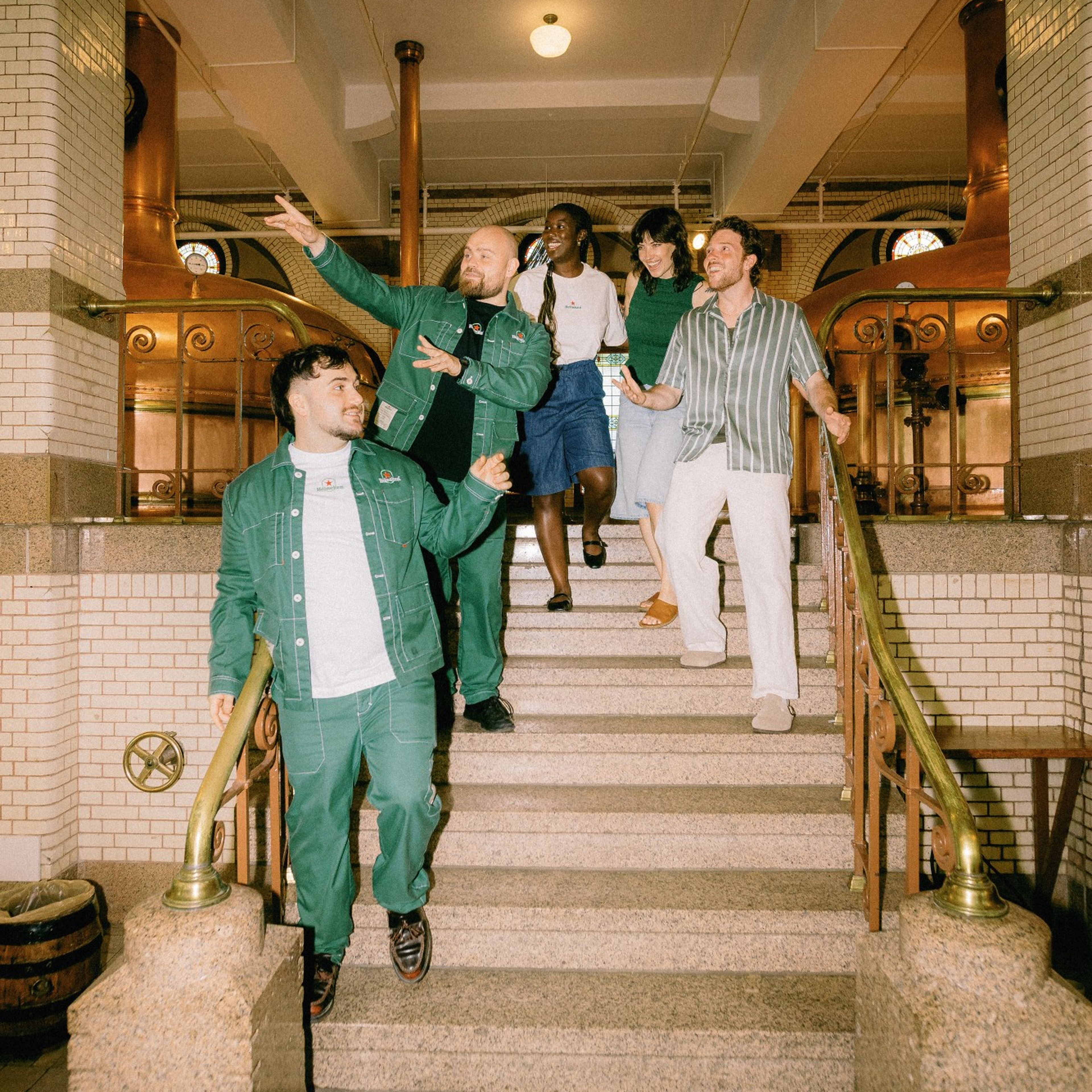 Five cheerful friends walk down grand stairs in an ornate building with copper fixtures, white tiles, and brass railings.