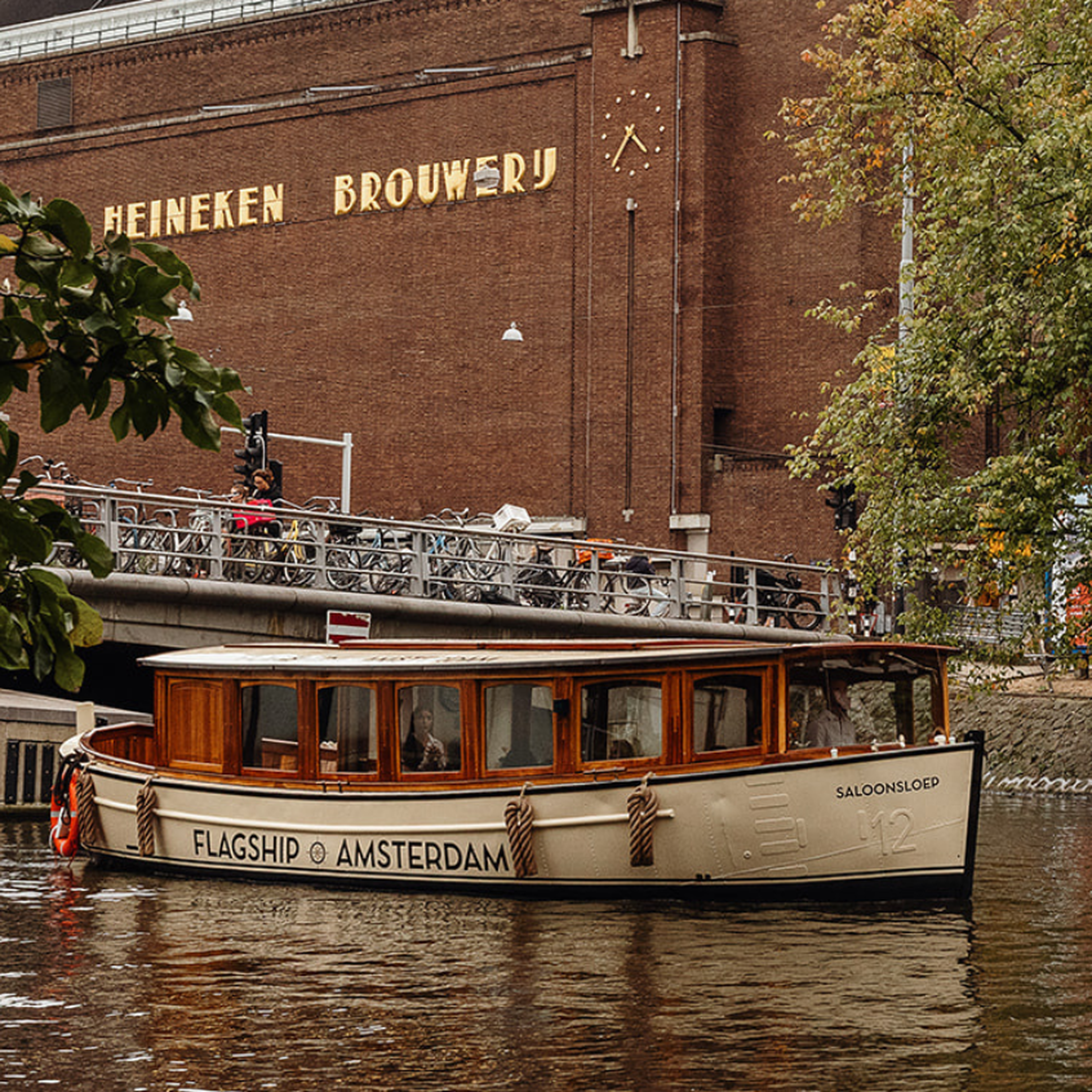Boat labeled "Flagship Amsterdam" on a canal near the Heineken Brewery, with trees and a bridge in the background.