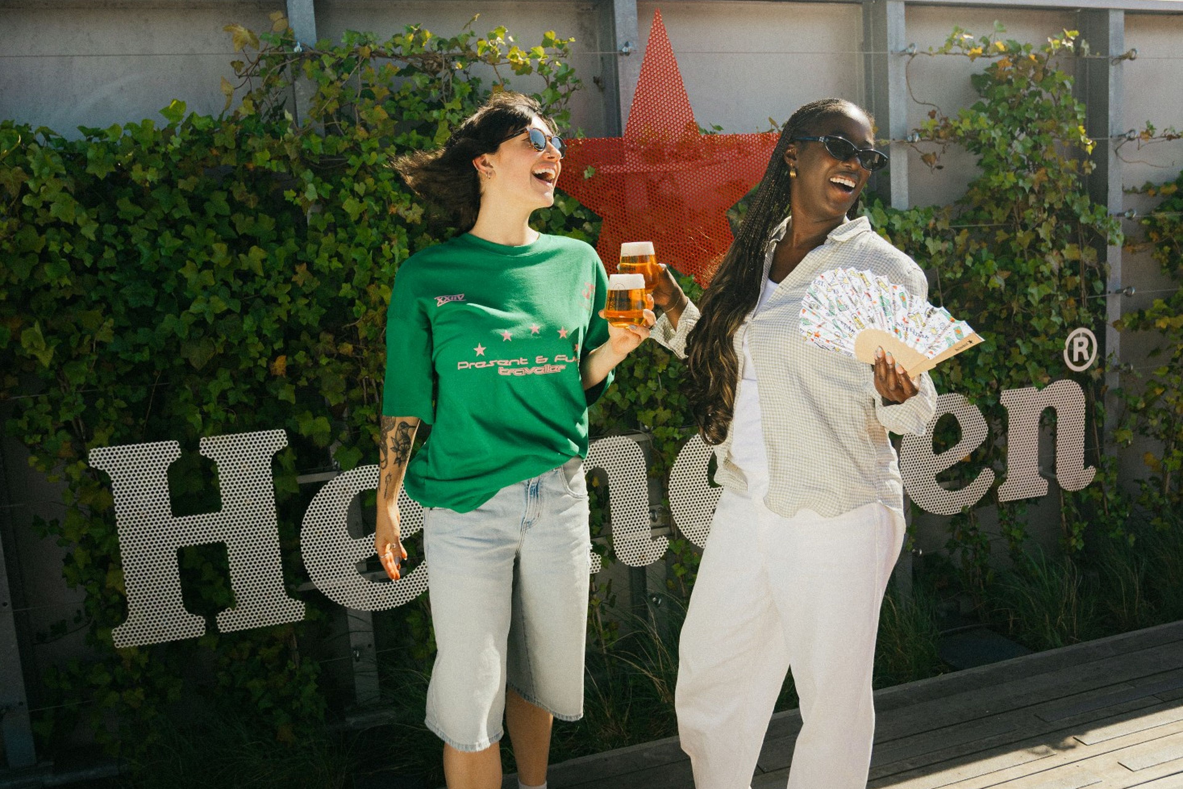 Two laughing women pose in front of a Heineken sign on a sunny rooftop deck, one holding a beer and the other a decorative fan.