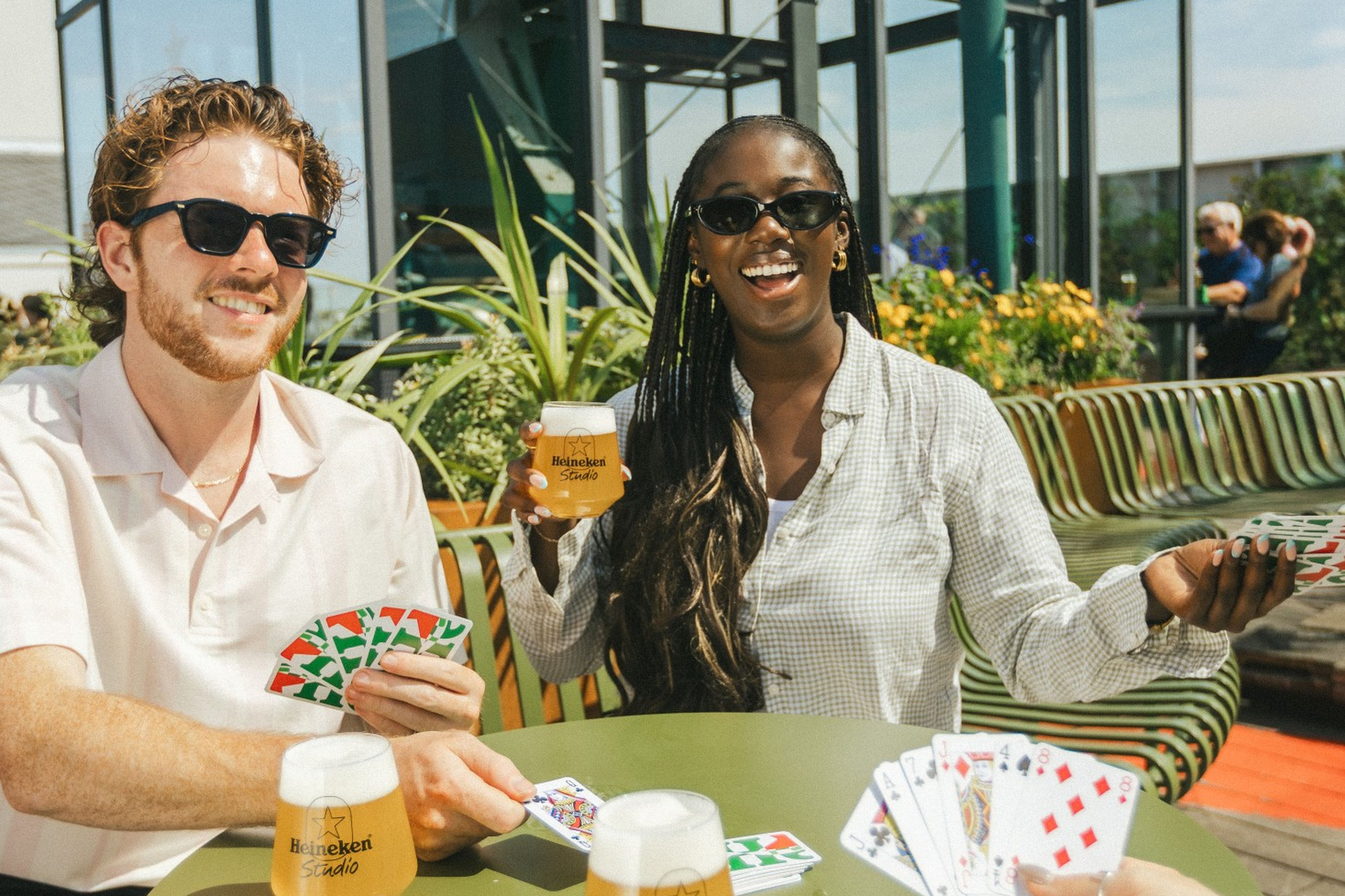 Two friends playing cards and enjoying Heineken beers at an outdoor rooftop terrace on a sunny day, laughing and wearing sunglasses.