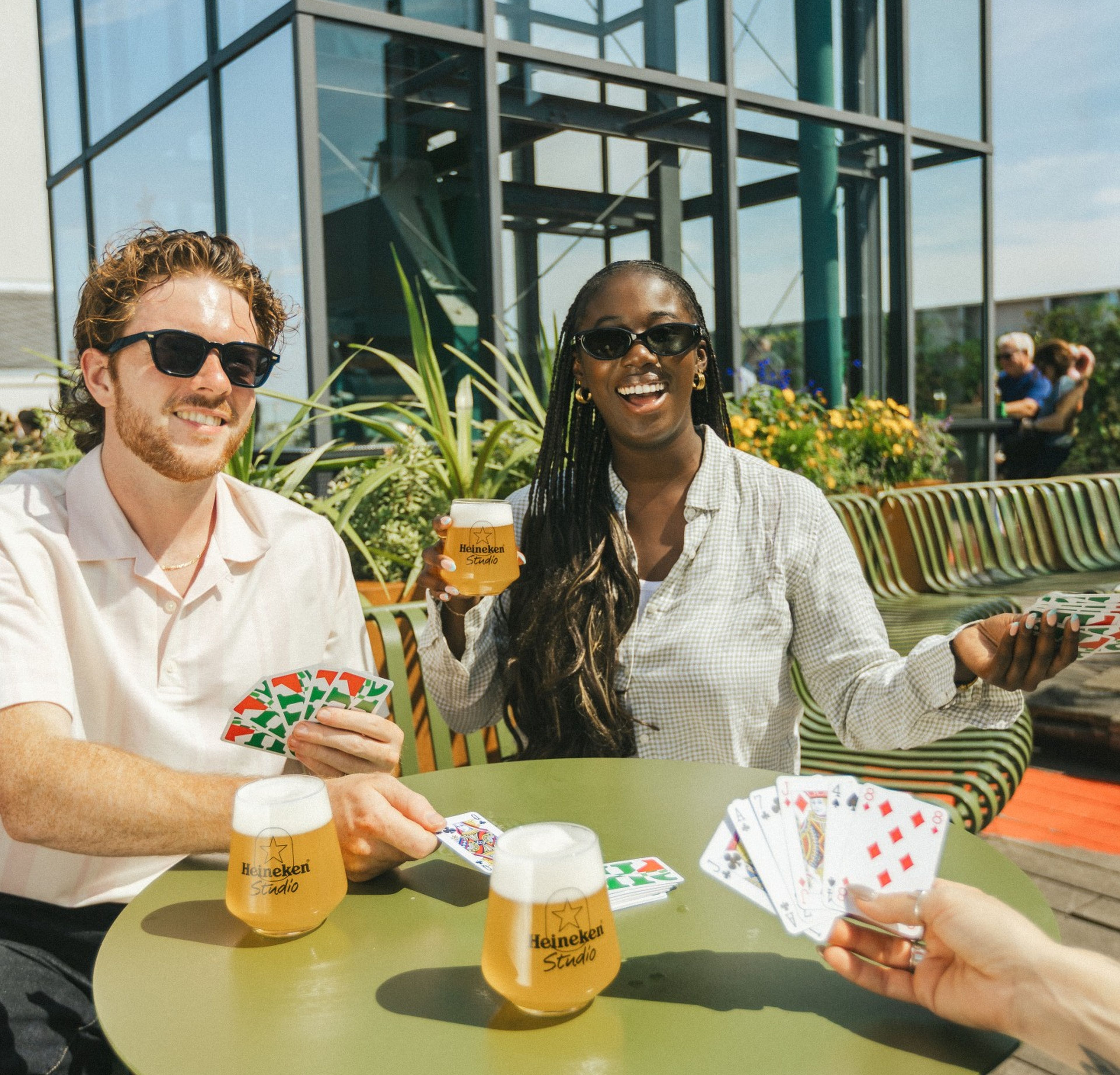 Two friends playing cards and enjoying Heineken beers at an outdoor rooftop terrace on a sunny day, laughing and wearing sunglasses.