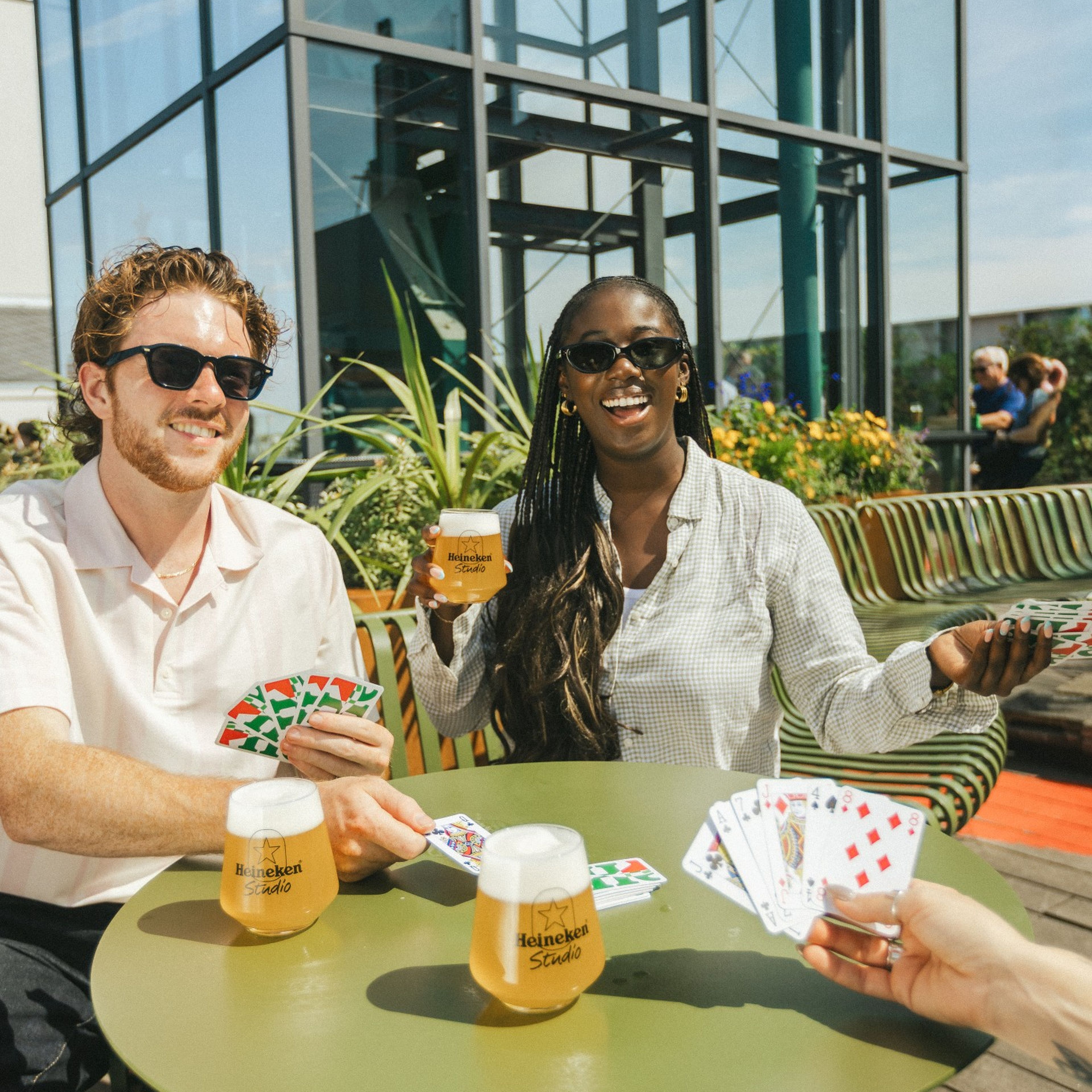 Two friends playing cards and enjoying Heineken beers at an outdoor rooftop terrace on a sunny day, laughing and wearing sunglasses.