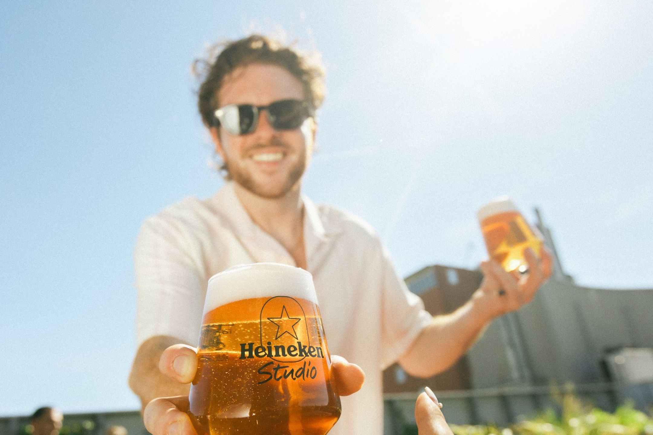 Smiling man in sunglasses handing a Heineken Studio branded beer glass to someone outdoors on a sunny day.