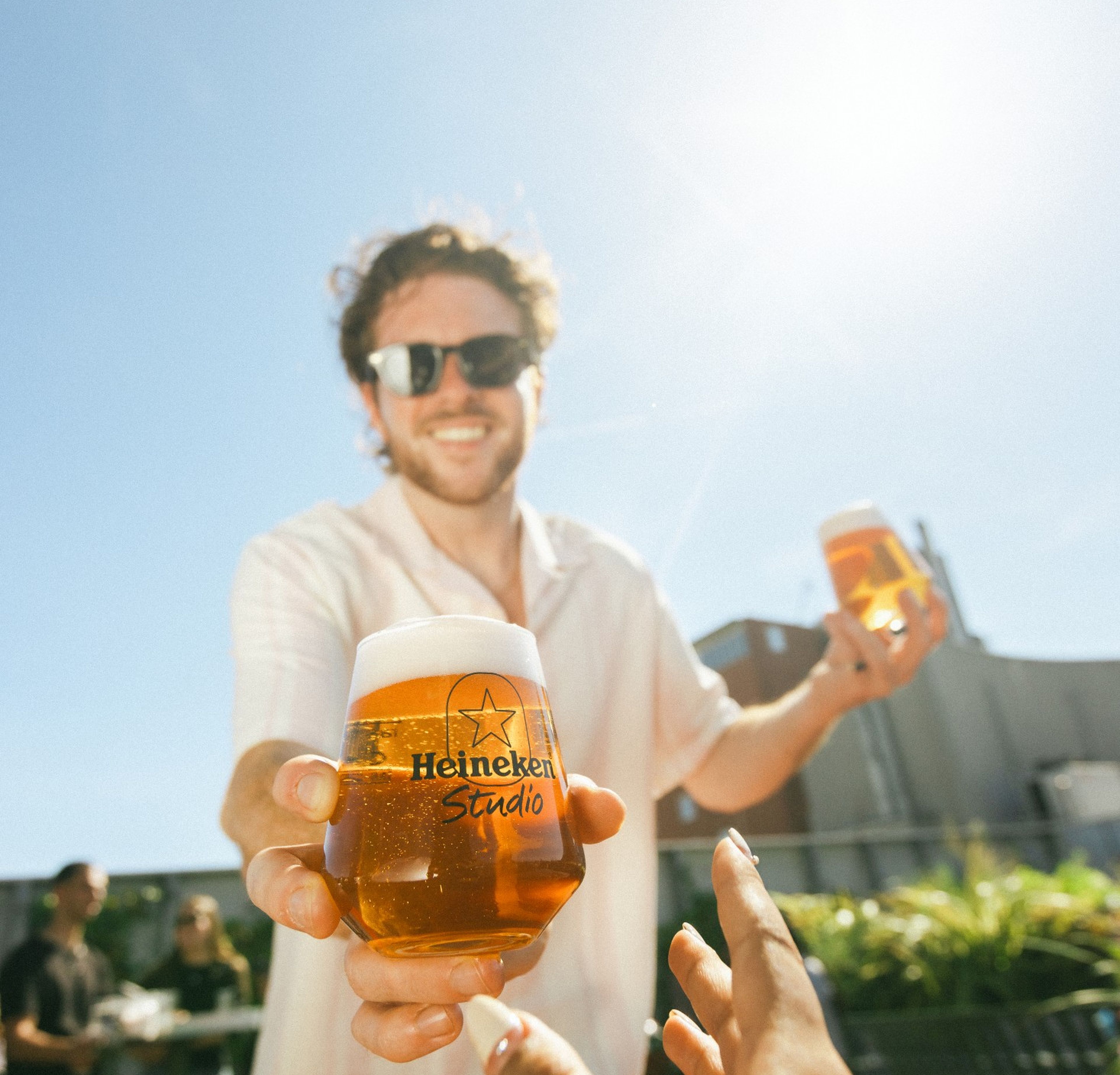 Smiling man in sunglasses handing a Heineken Studio branded beer glass to someone outdoors on a sunny day.