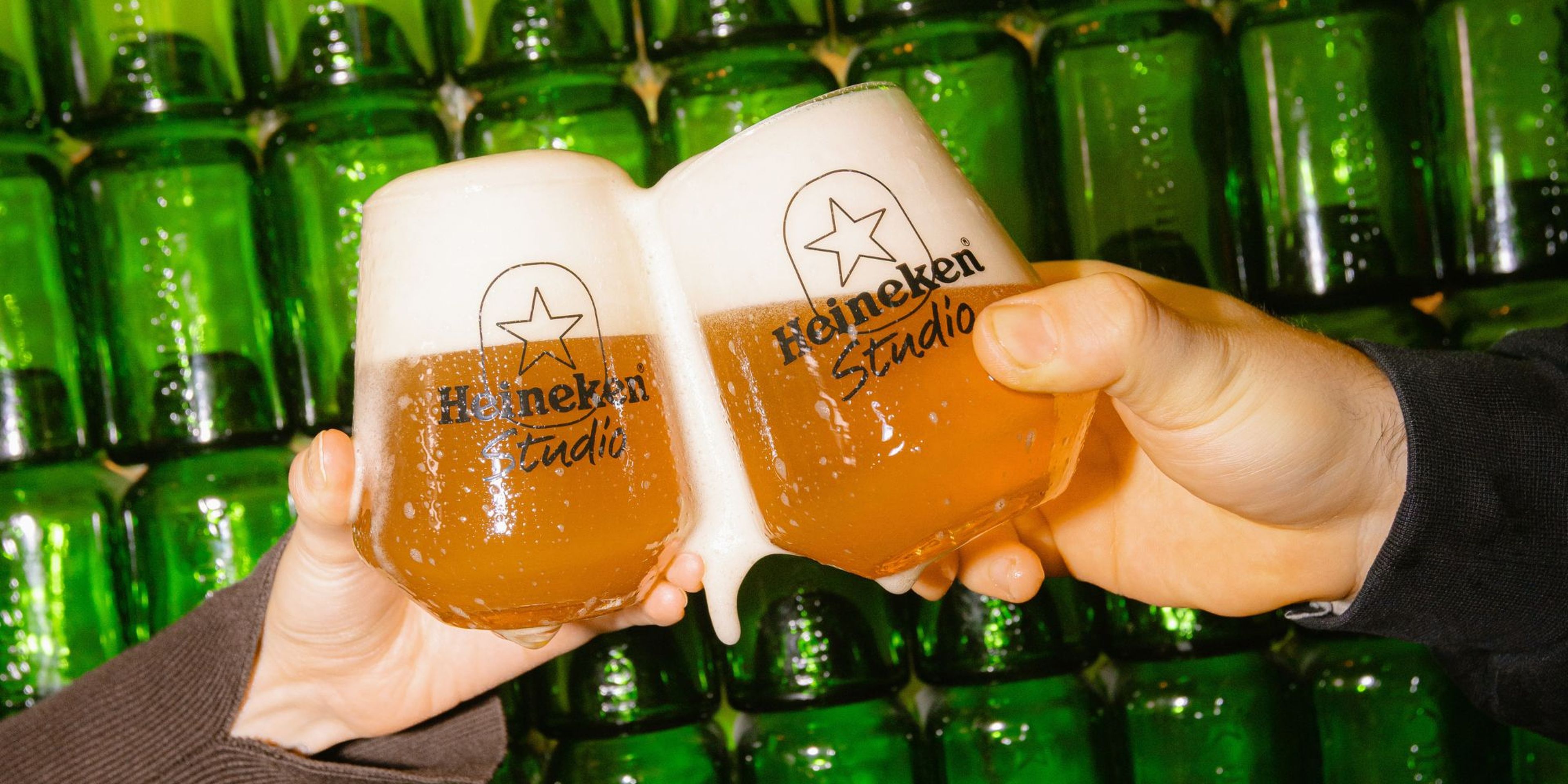 Two hands clinking Heineken glasses filled with beer, against a backdrop of green bottles.