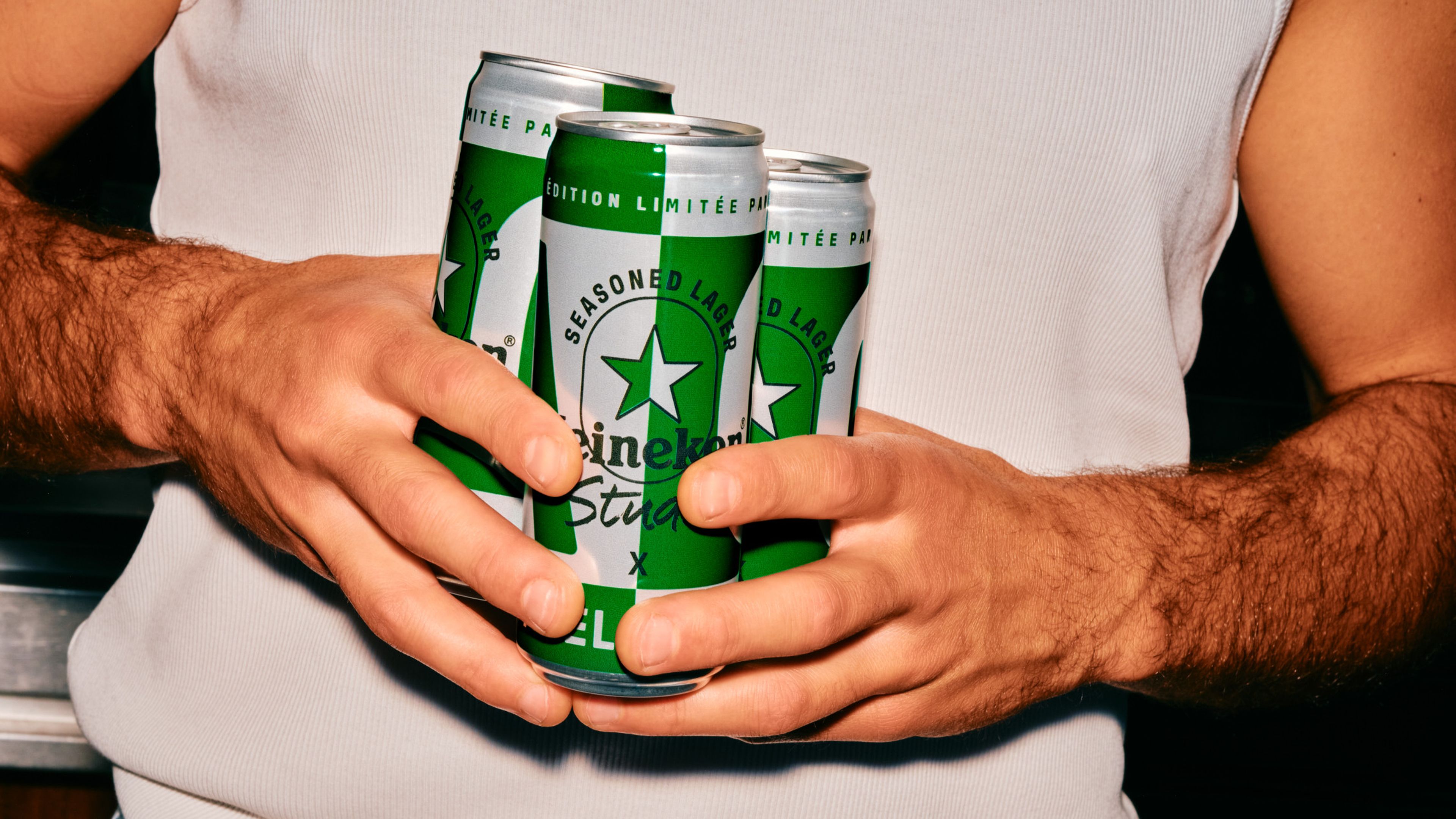 A person in a white tank top holds four green cans of Heineken beer, showcasing the brand's logo and design.