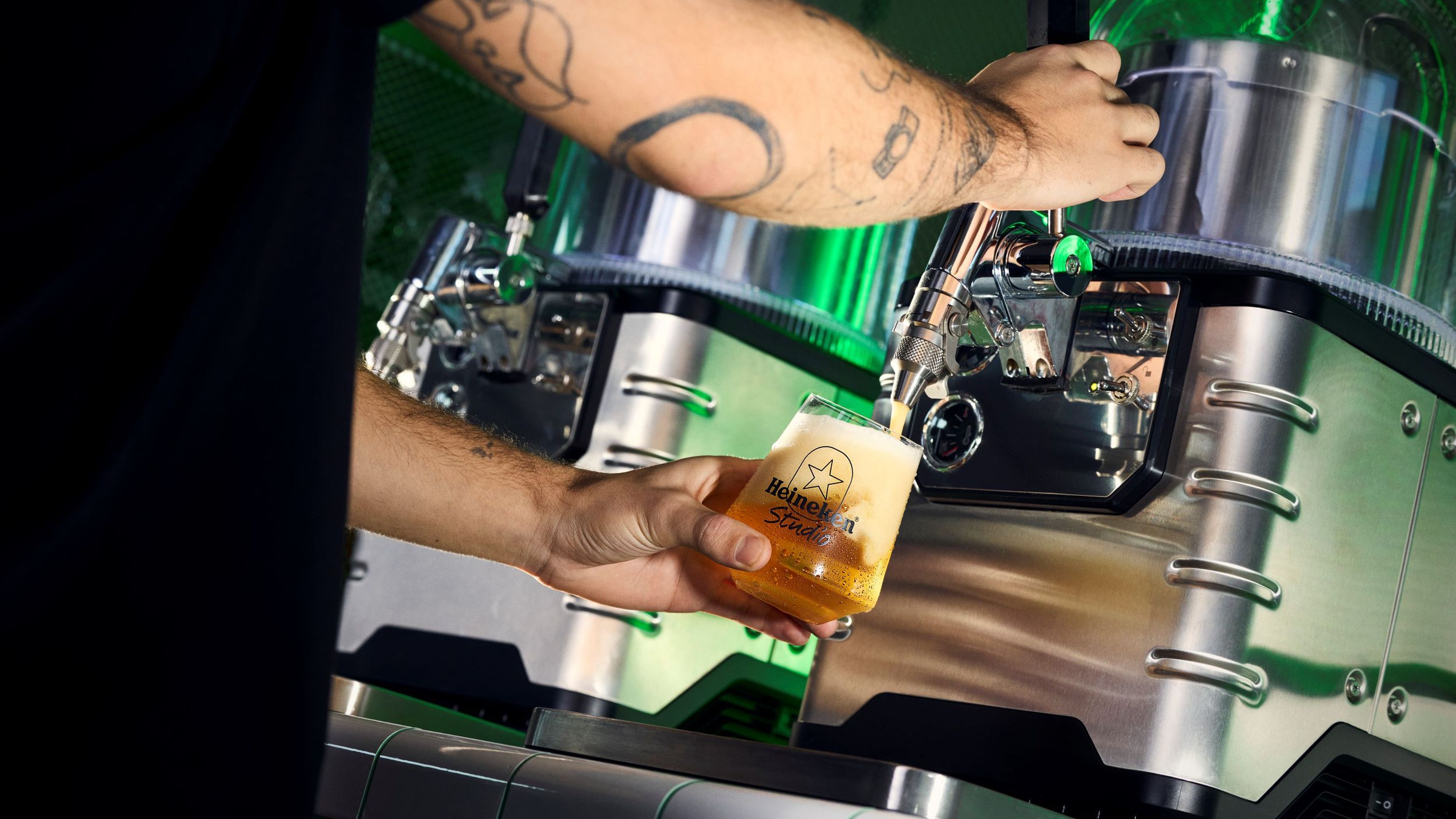 Person with tattoos pours beer into a Heineken Studio glass from a modern dispenser, with green neon lights in the background.