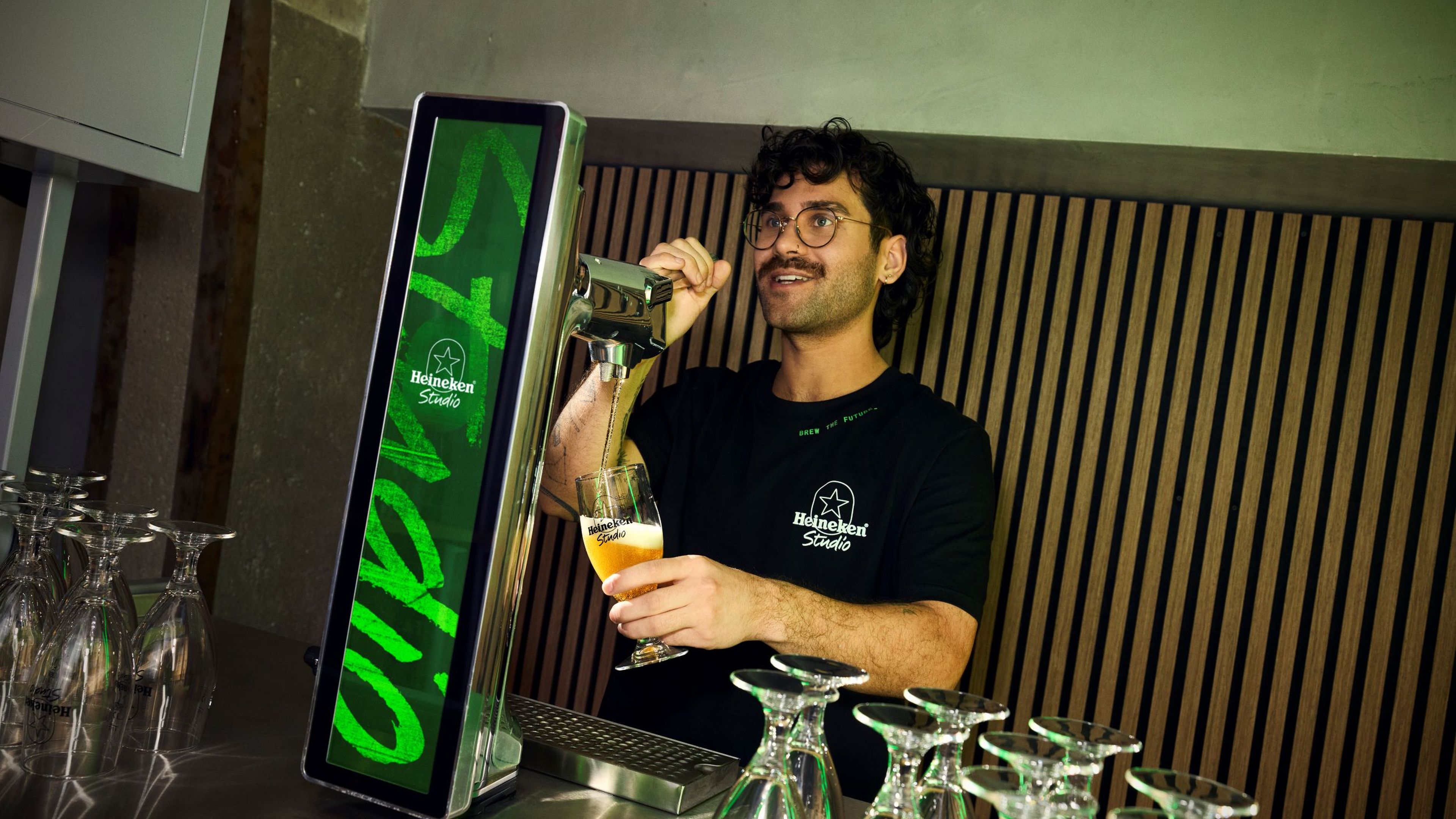 A man pours a beer from a Heineken tap, surrounded by upside-down glasses on a bar counter.