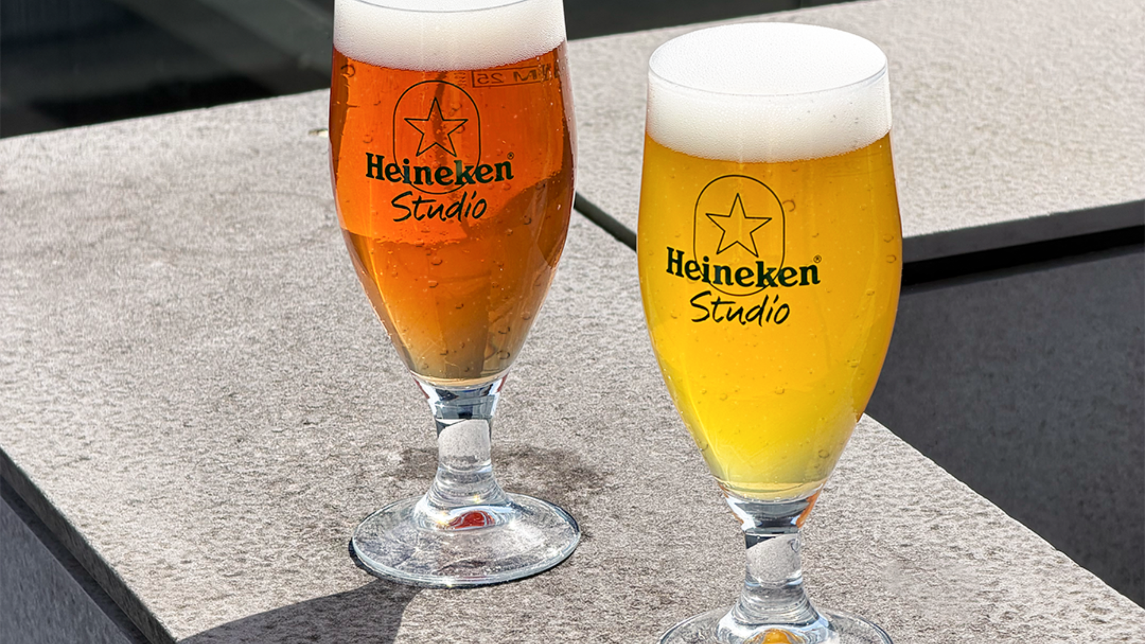 Two glasses of Heineken Studio beer, one amber and one golden, with foamy heads, placed on a stone surface outdoors.