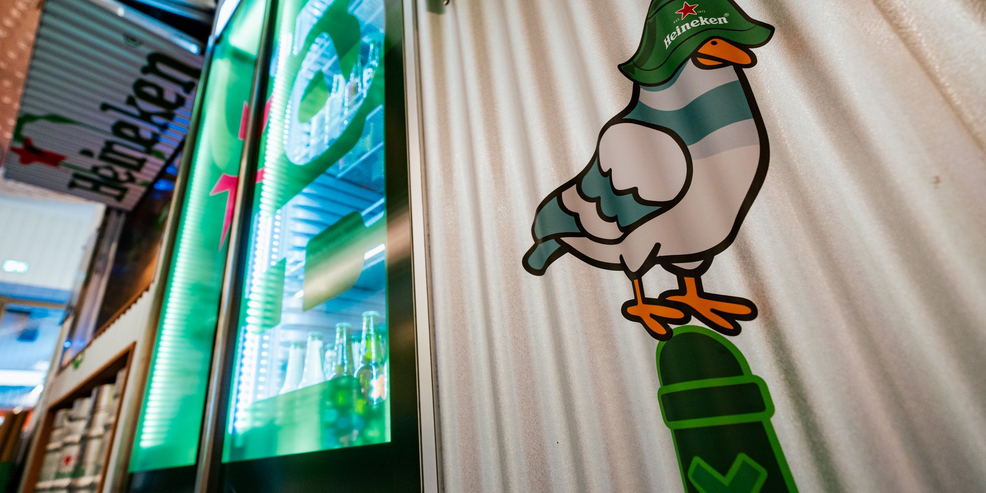 Cartoon bird wearing a Heineken hat on a wall, next to illuminated Heineken fridges displaying bottles inside a store.