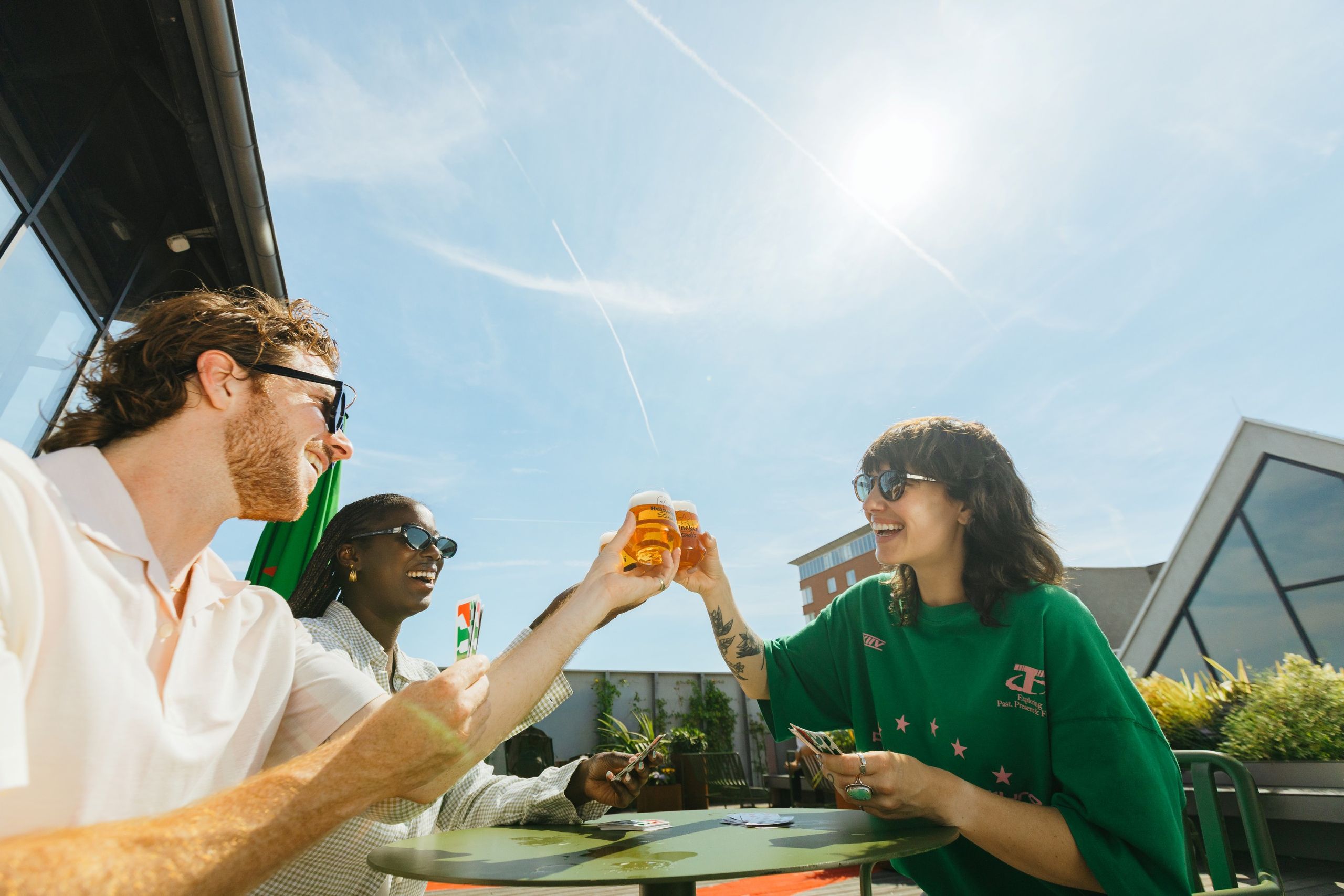 Three friends laughing and toasting with beer glasses on a sunny rooftop terrace, playing cards on a green table.