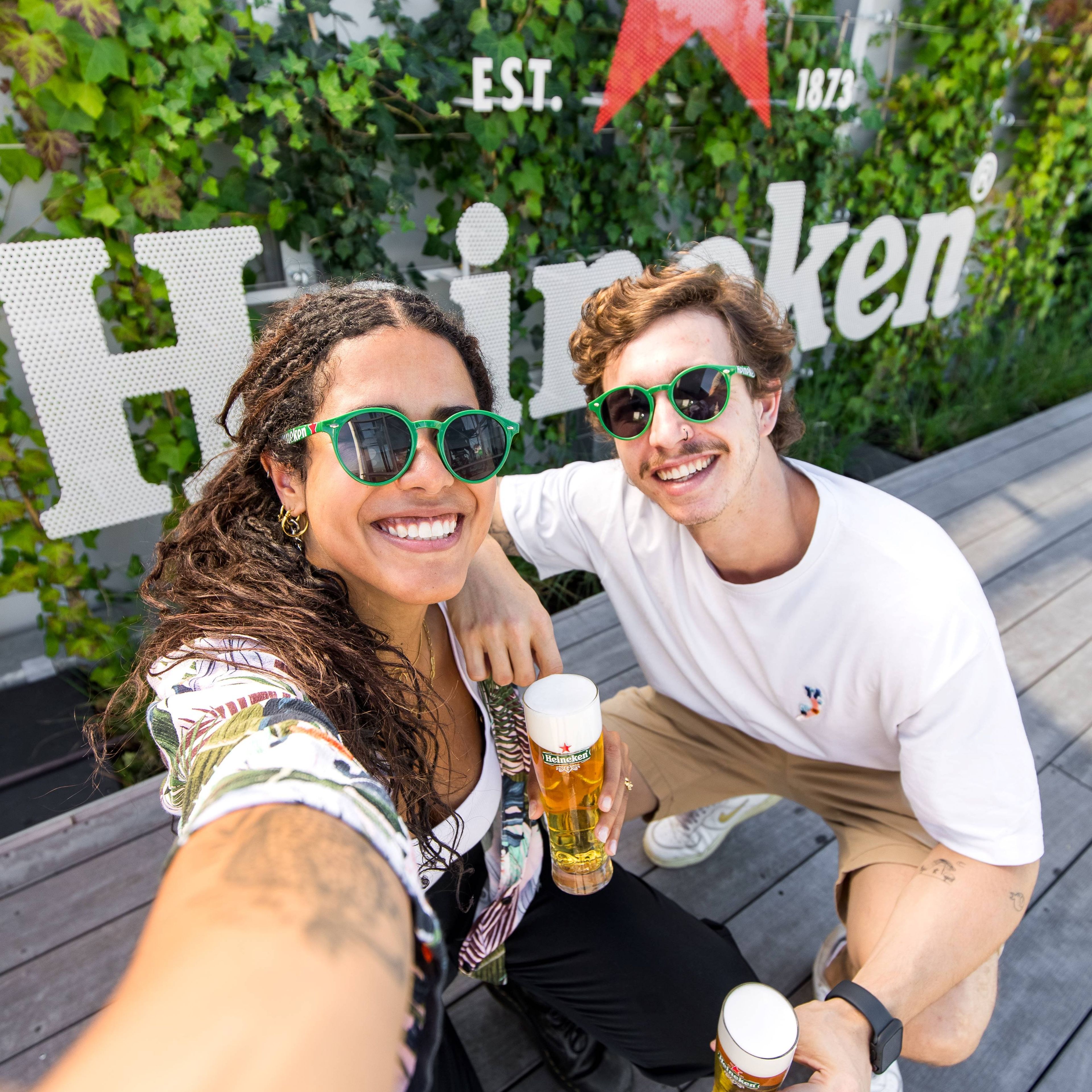 Two people enjoying drinks on a rooftop with plants and a Heineken sign in the background.