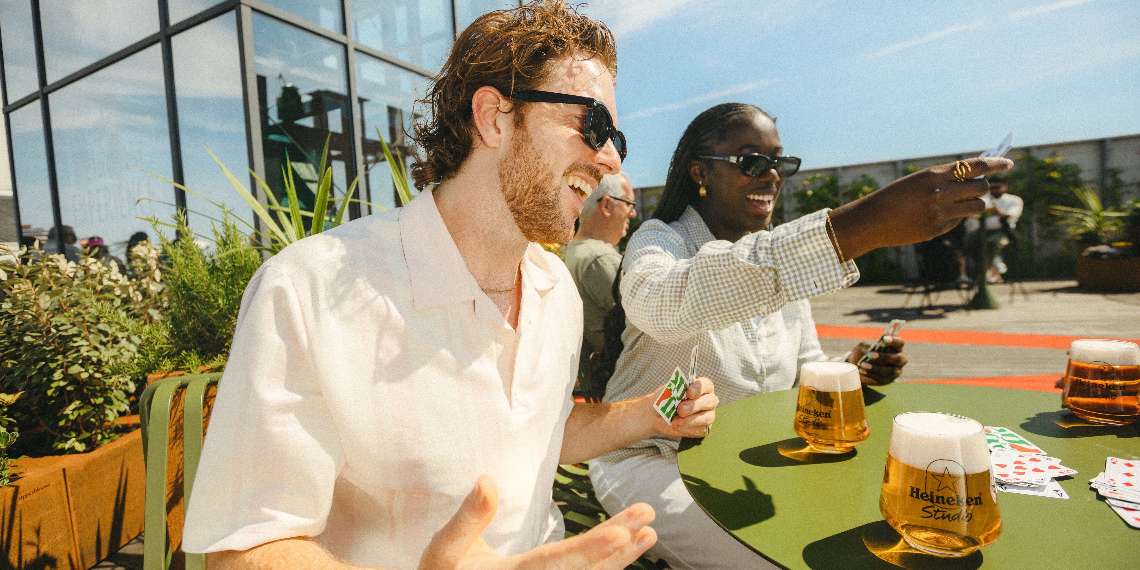 Two friends enjoying Heineken beer and playing cards on a sunny outdoor rooftop terrace on a bright summer day.