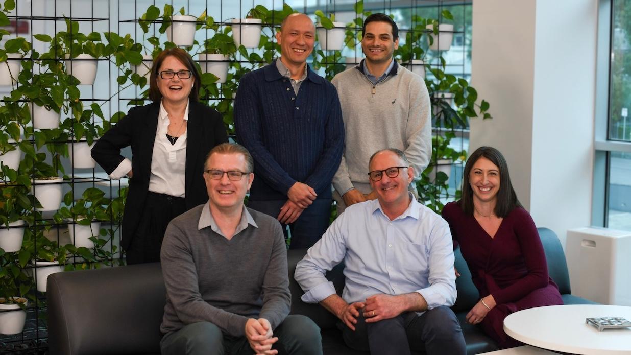 Six smiling professionals, three standing and three seated, in a modern office with a green wall of potted plants.