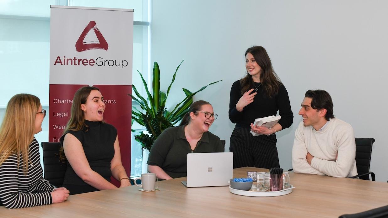 Five colleagues from Aintree Group, four seated and one standing, engaging in a lively discussion, all laughing and smiling in an office meeting.