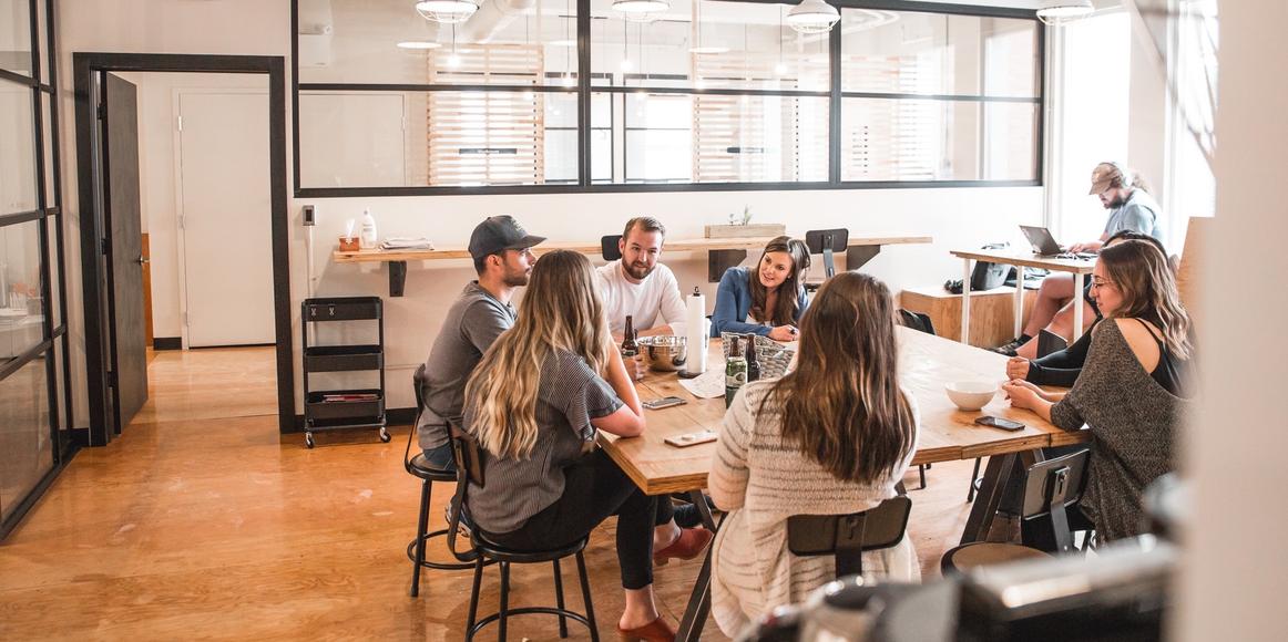 A group of 7 people sitting around an office table in discussion. There is someone in the background working with headphones on.