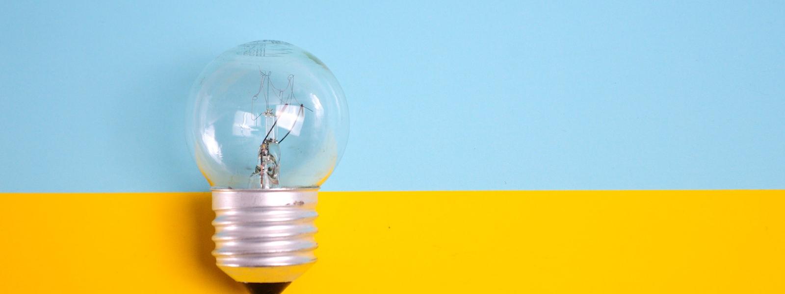 A light bulb in front of a wall that is painted light blue on the top and bright yellow on the bottom.