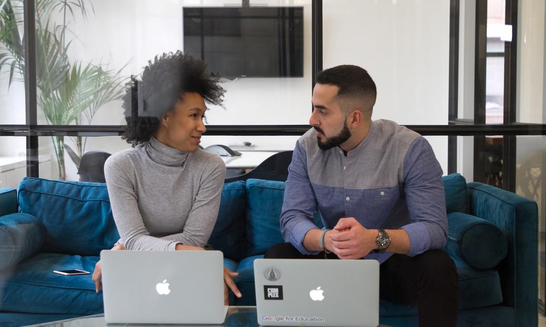 Two people sitting next to each other on a sofa in discussion, with their laptops on the table in front of them.