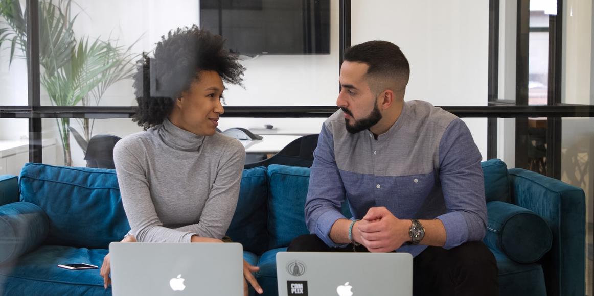 Two people sitting next to each other on a sofa in discussion, with their laptops on the table in front of them.