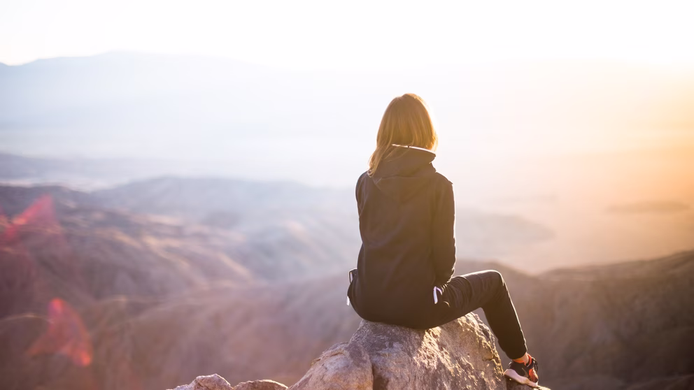 A person is sitting with their back to the camera on a rock and looking out over an expansive view of a rock formation.