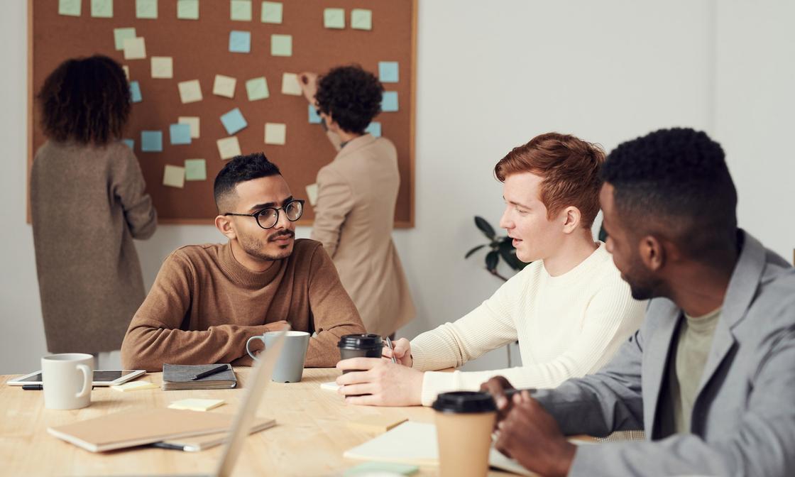 3 people sitting around a desk making business decisions, with 2 others standing behind them in front of a pinboard covered in sticky notes.