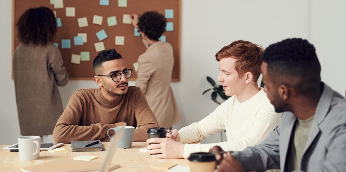 3 people sitting around a desk making business decisions, with 2 others standing behind them in front of a pinboard covered in sticky notes.