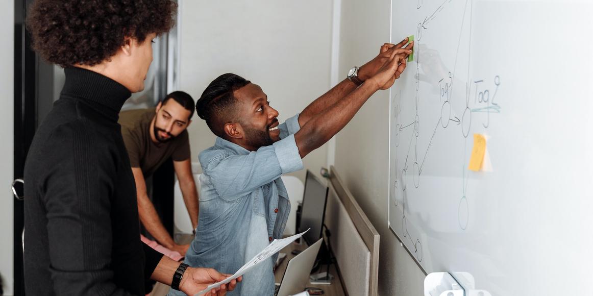 3 people are working together, in conversation, as 1 person is reaching to stick a Post-It note on a whiteboard.