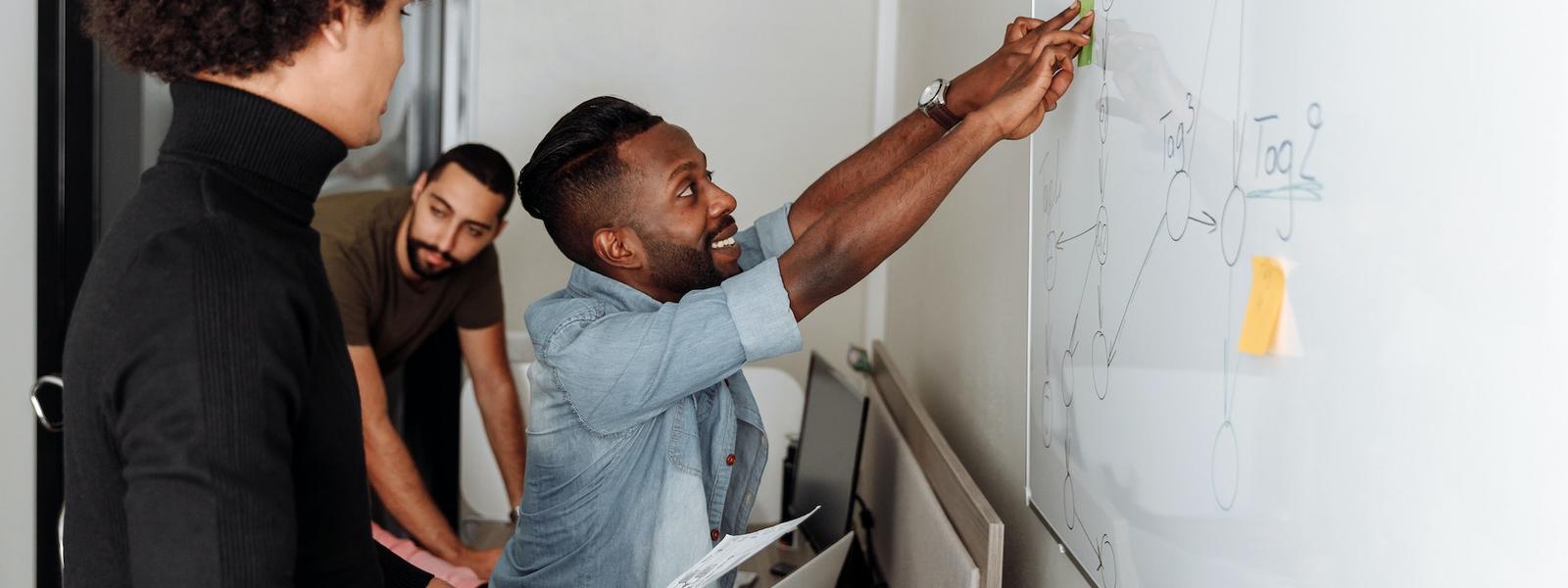 3 people are working together, in conversation, as 1 person is reaching to stick a Post-It note on a whiteboard.