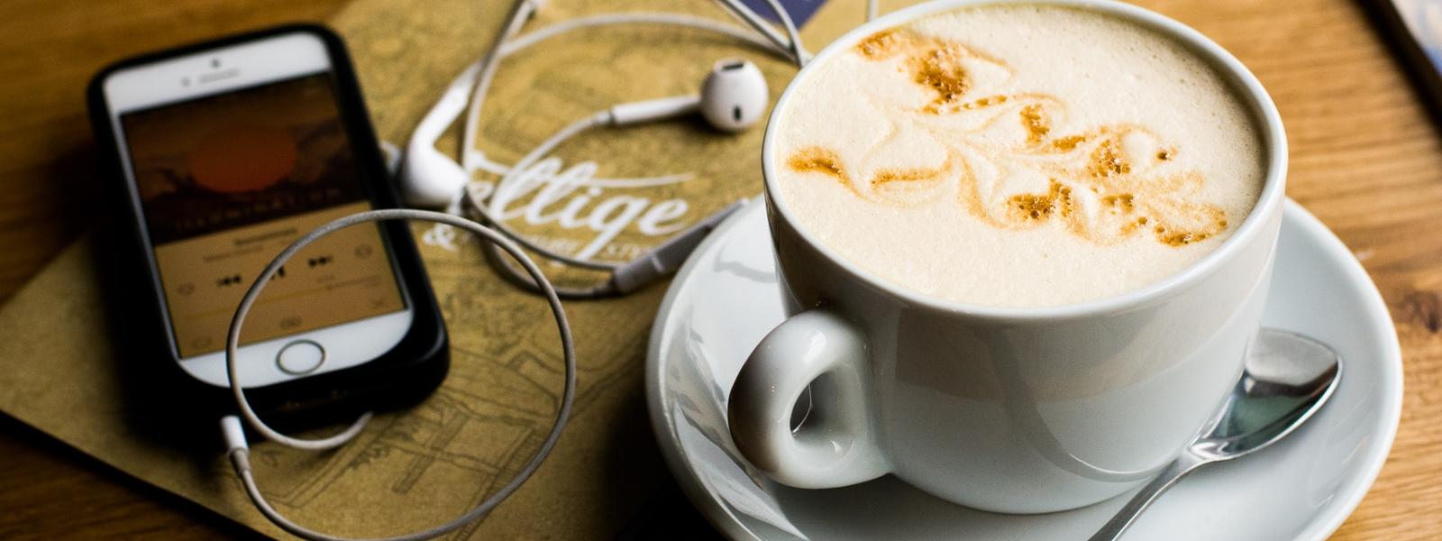 On a timber table is a phone playing a podcast with headphones plugged in, next to a coffee.