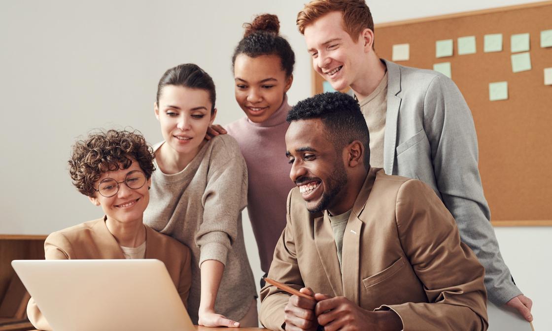 5 young professionals looking at a laptop screen and smiling. There is a pegboard behind them with green stickie notes.