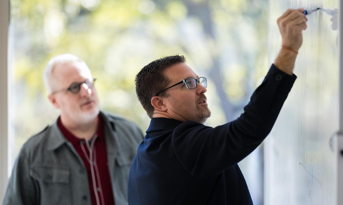 Ian Vacin and Jason Blumer stand at a whiteboard with Ian writing while Jason watches attentively during a planning discussion in a bright office.