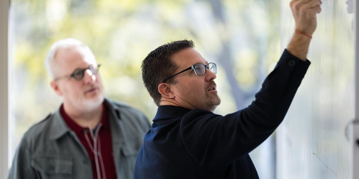 Ian Vacin and Jason Blumer stand at a whiteboard with Ian writing while Jason watches attentively during a planning discussion in a bright office.