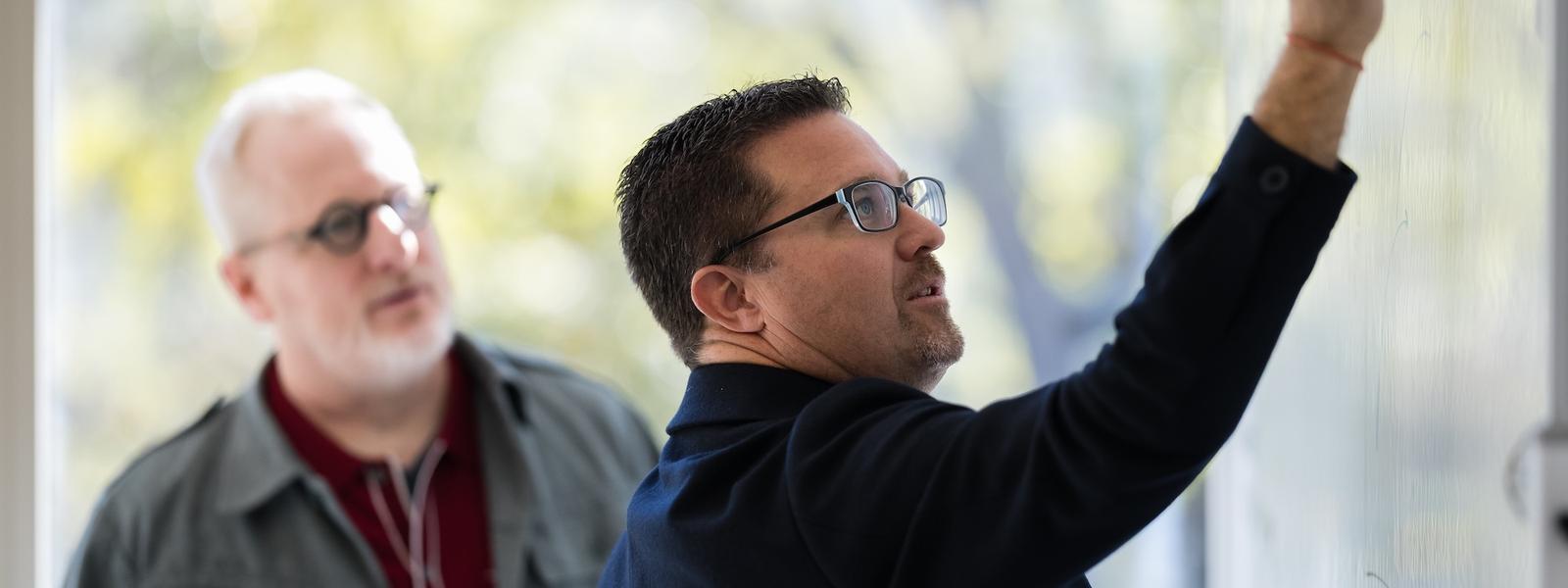 Ian Vacin and Jason Blumer stand at a whiteboard with Ian writing while Jason watches attentively during a planning discussion in a bright office.