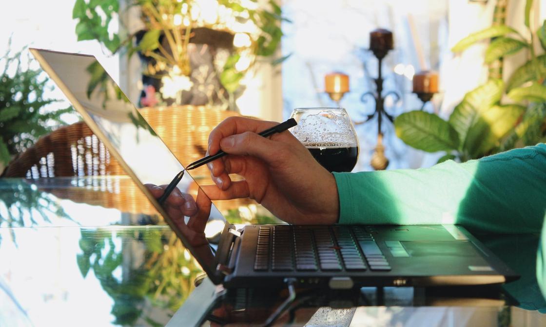 An arm with a green shirt sleeve holding a digital pen to a laptop with a beer next to it in a well-lit room filled with plants.