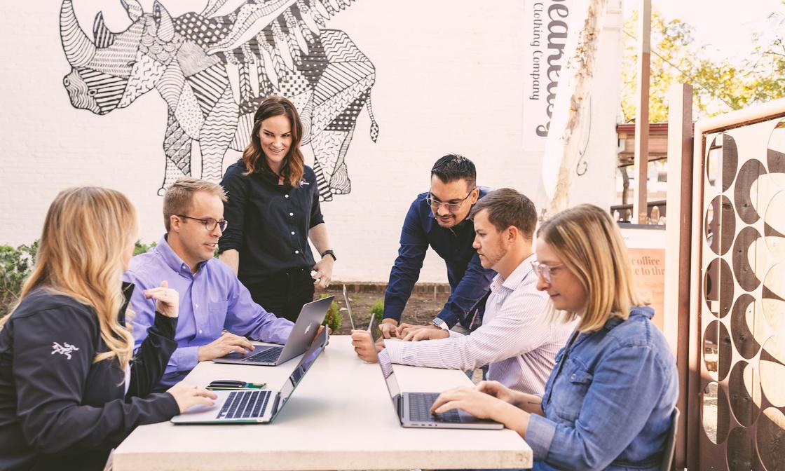 6 Karbon staff members surrounding a table outdoors. 4 of them are sitting at the table, working from laptops. The other 2 are standing, looking over the others and engaged in conversations.