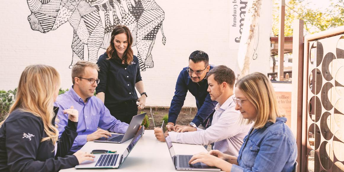 6 Karbon staff members surrounding a table outdoors. 4 of them are sitting at the table, working from laptops. The other 2 are standing, looking over the others and engaged in conversations.